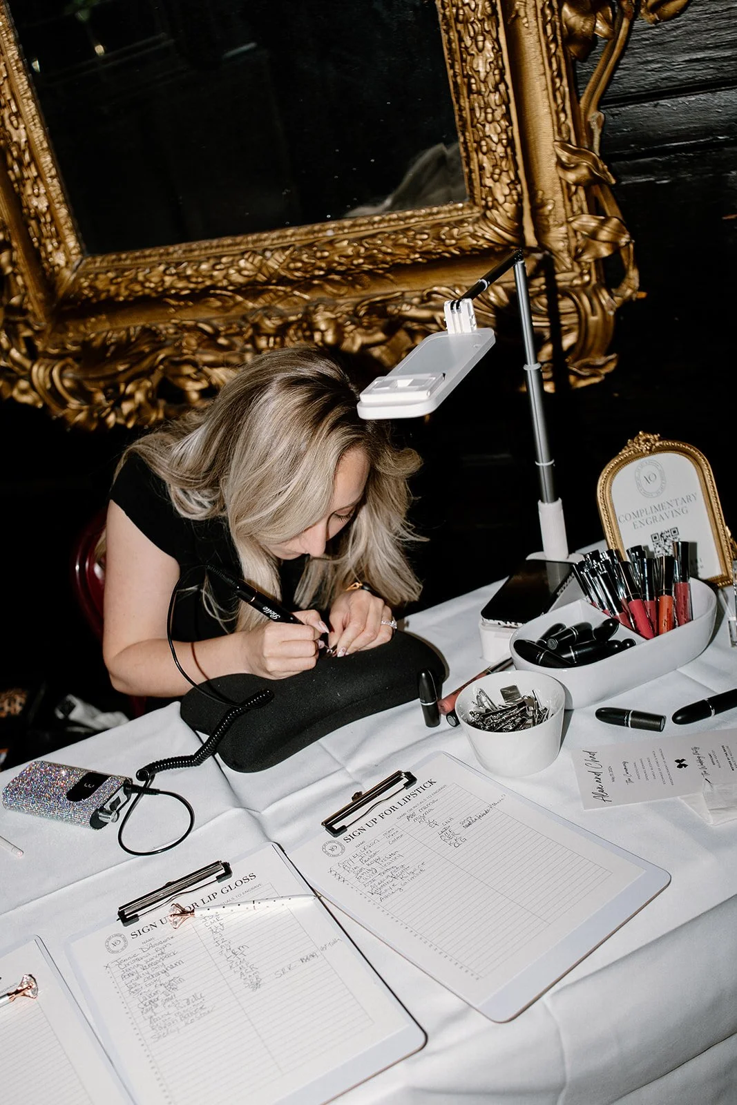 A woman getting a lip tattoo at a table with various tattoo equipment, including lip glosses and sign-up sheets, against a ornate gold-framed mirror backdrop.