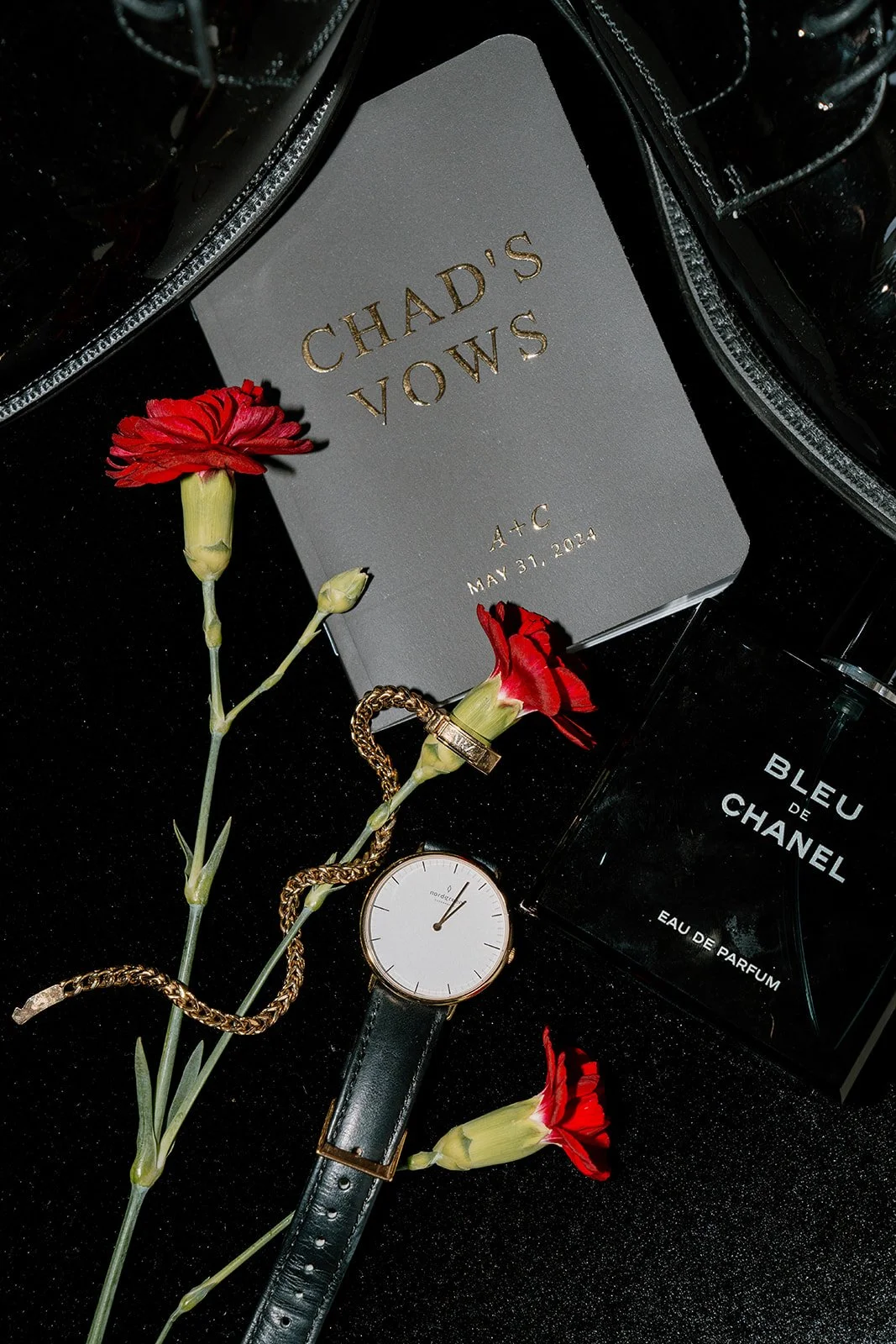 A gray notebook with the words "Chad's Vows," a gold bracelet, a black wristwatch with a white face and black strap, a bottle of Bleu de Chanel perfume, and three red flowers on a black surface.