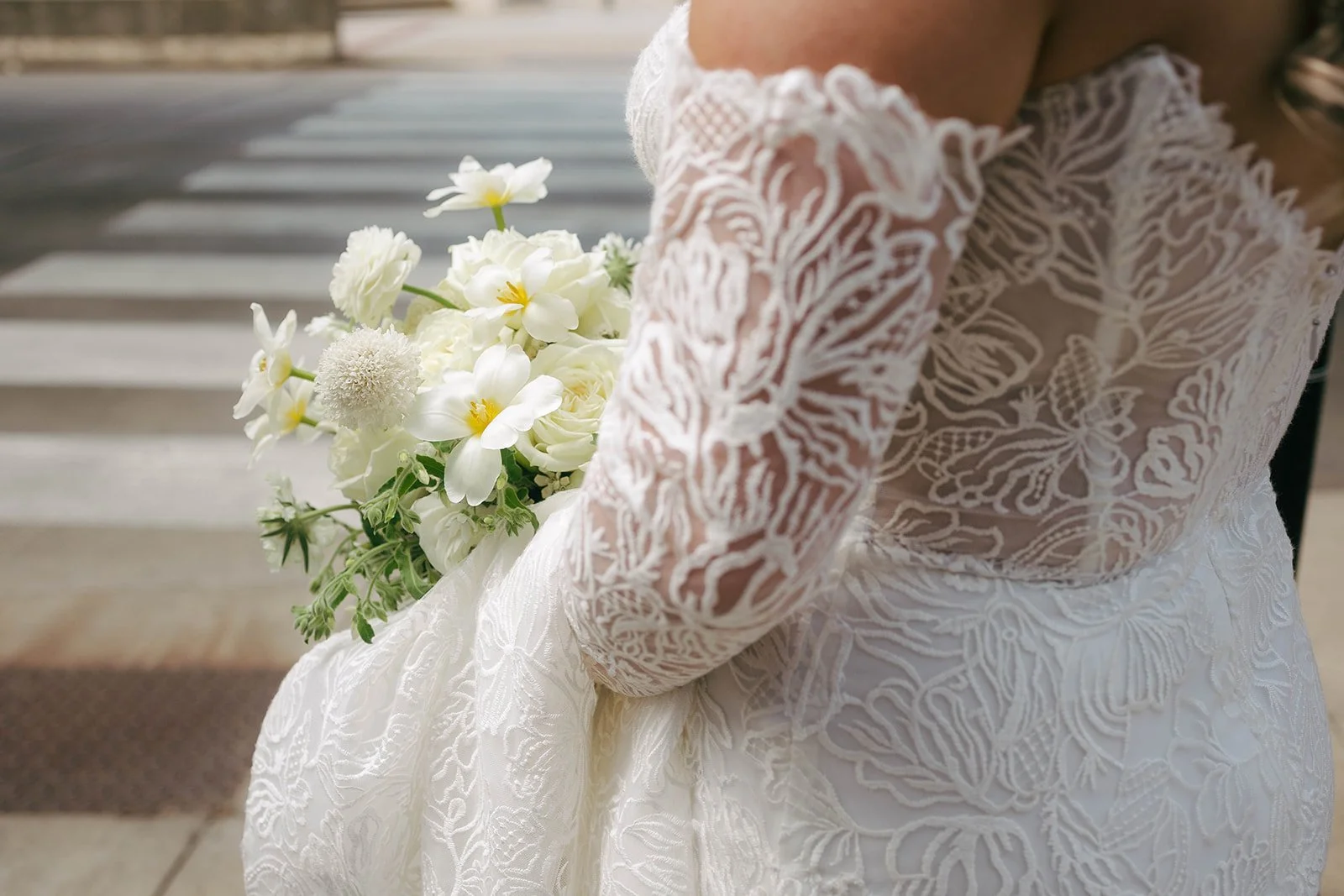 Bride wearing a lace wedding dress holding a bouquet of white flowers outside.