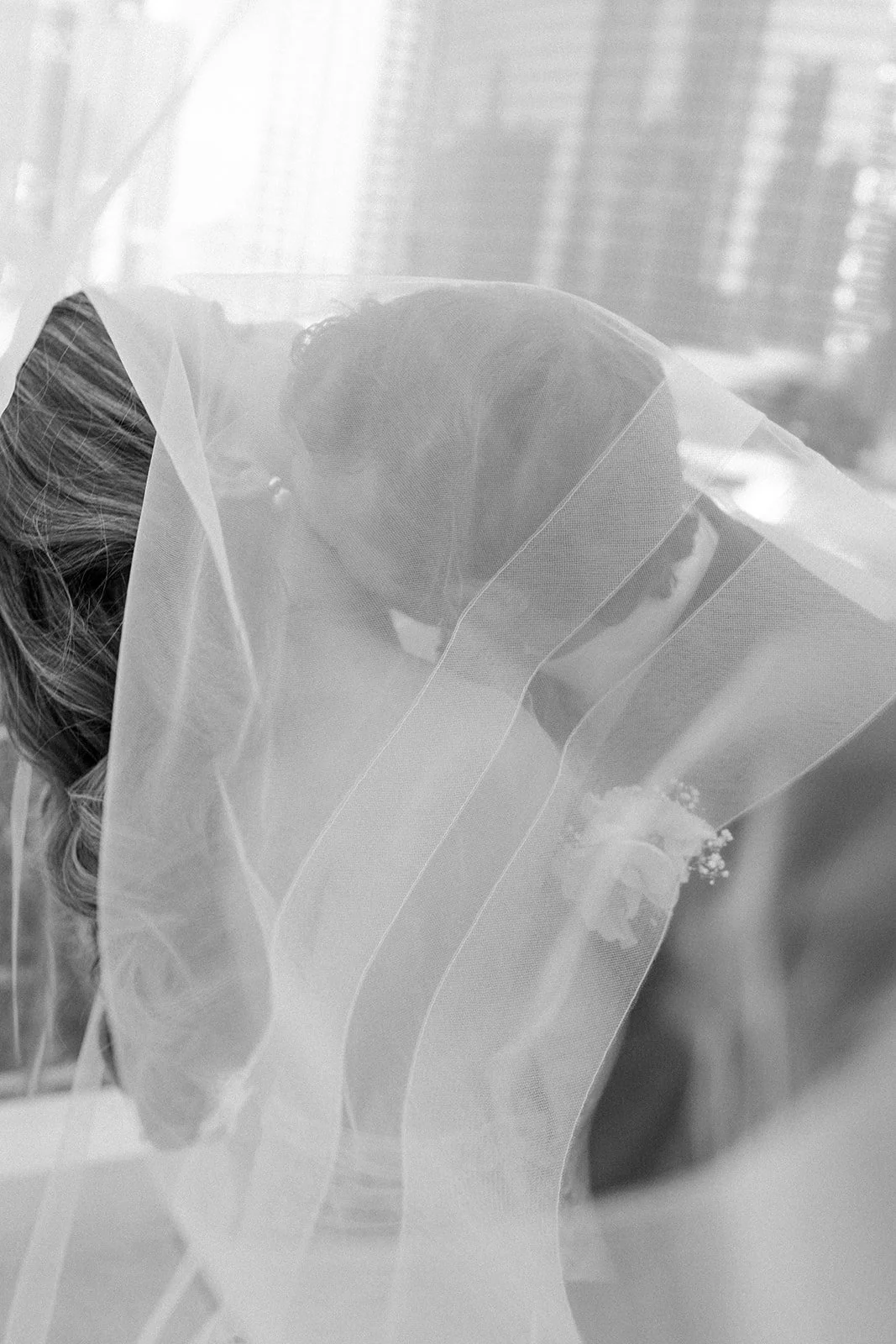 A woman, possibly a bride, with curly hair and a veil over her face, is leaning forward and appears to be kissing or resting her forehead on a bouquet of flowers.