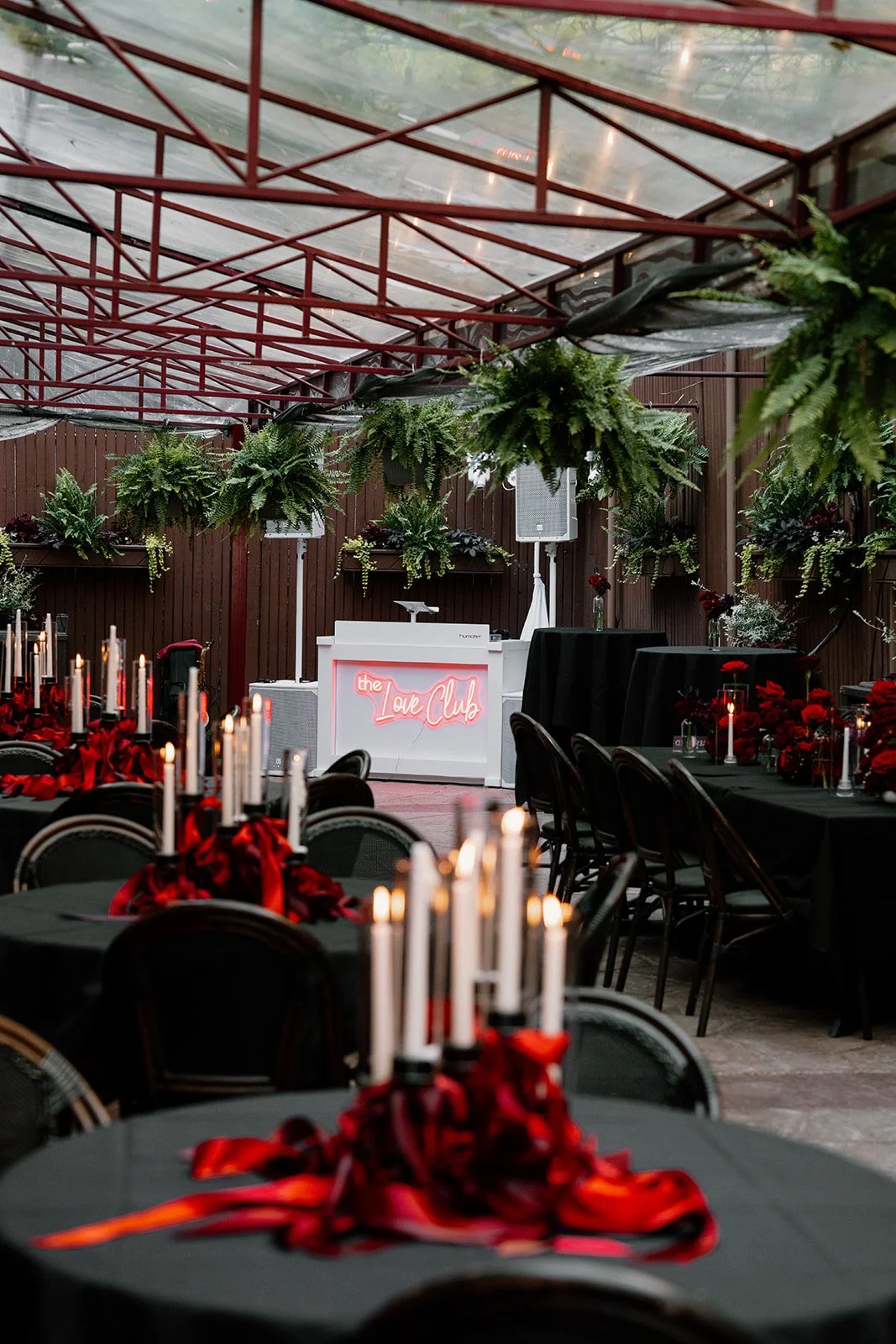 Event space decorated with black tablecloths, red and black floral centerpieces, and tall white candles. Hanging lush greenery and ferns decorate the ceiling. A white DJ booth with a neon sign reads 'the Love Club'.
