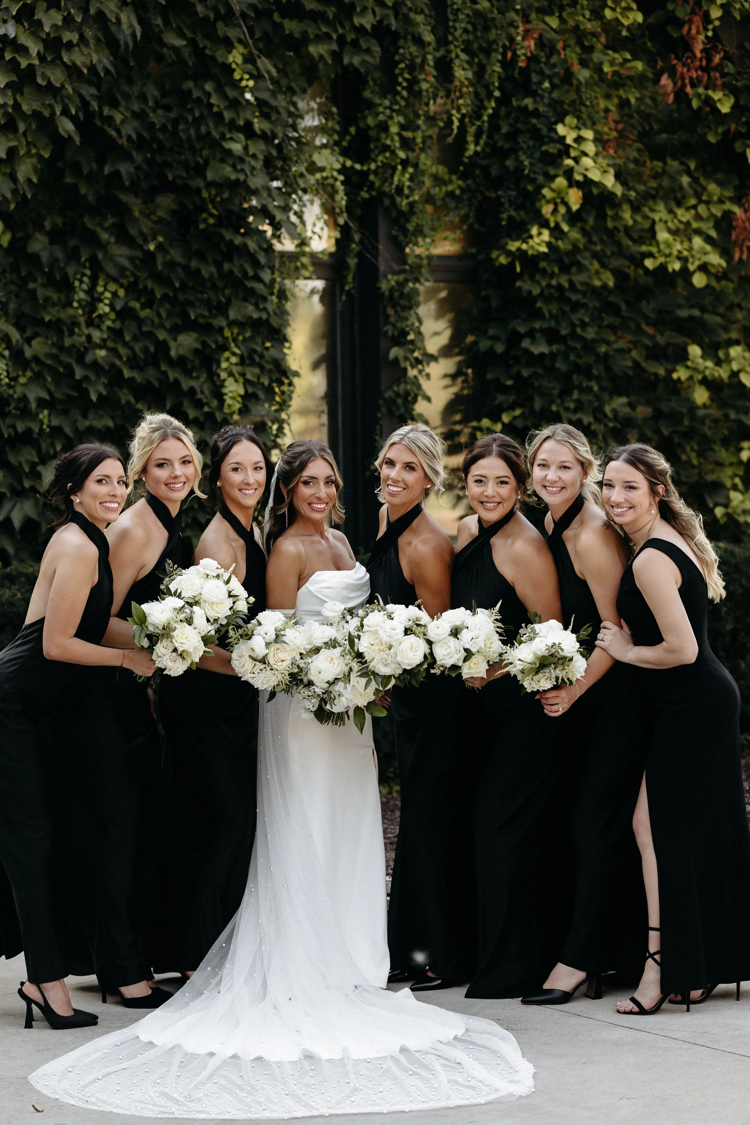 Bride in a white wedding gown with bridesmaids in black dresses, holding white bouquets, standing outdoors in front of a large leafy wall.