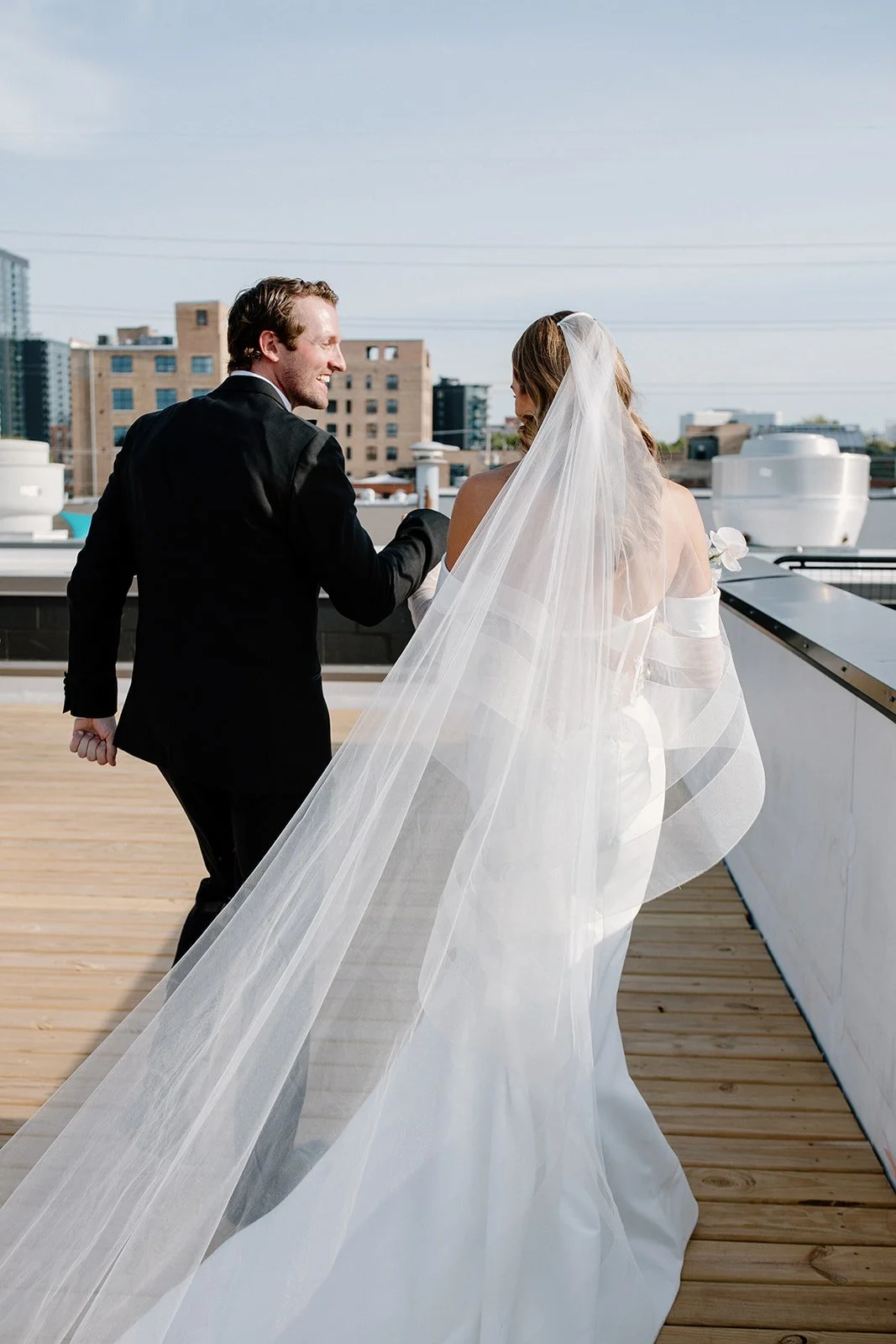 A happy newlywed couple on a rooftop, with the groom in a black suit holding the bride's hand, who is wearing a white wedding dress and a veil, against an urban cityscape background.