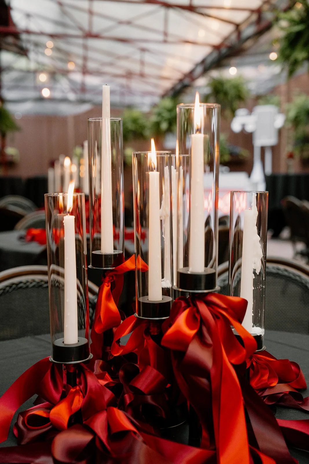Decorative centerpiece with tall white candles in glass holders and red ribbons, set on a dark table at an outdoor event under a metal canopy.