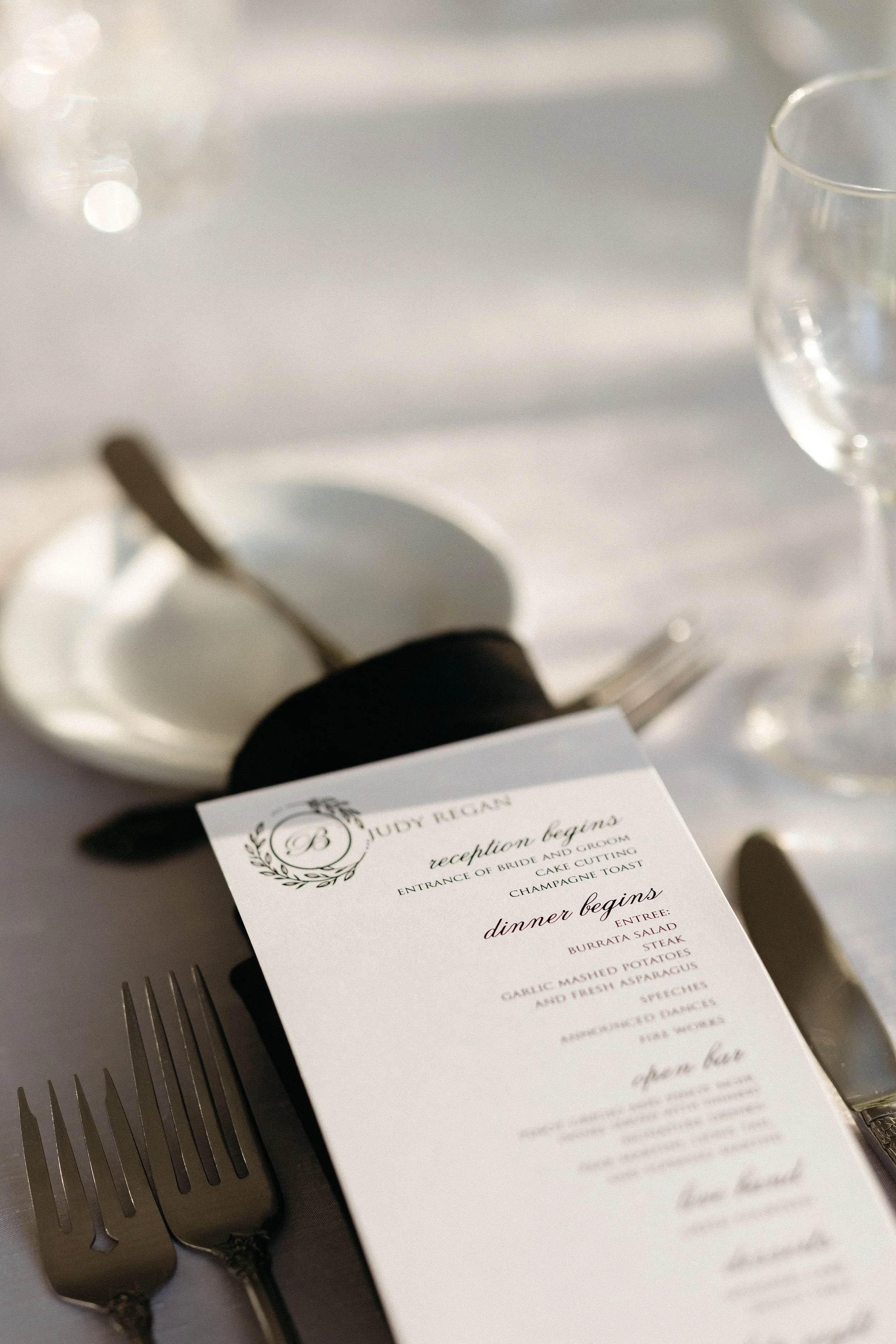 Wedding reception table setting with a menu, forks, a knife, an empty plate with a spoon, and a glass of water.