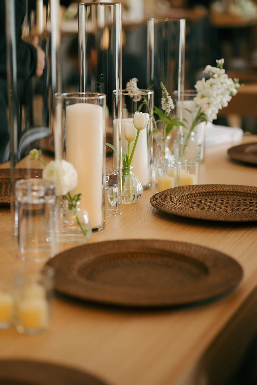 A wooden dining table decorated with white and cream-colored flowers in glass vases, tall candles, and woven brown placemats.
