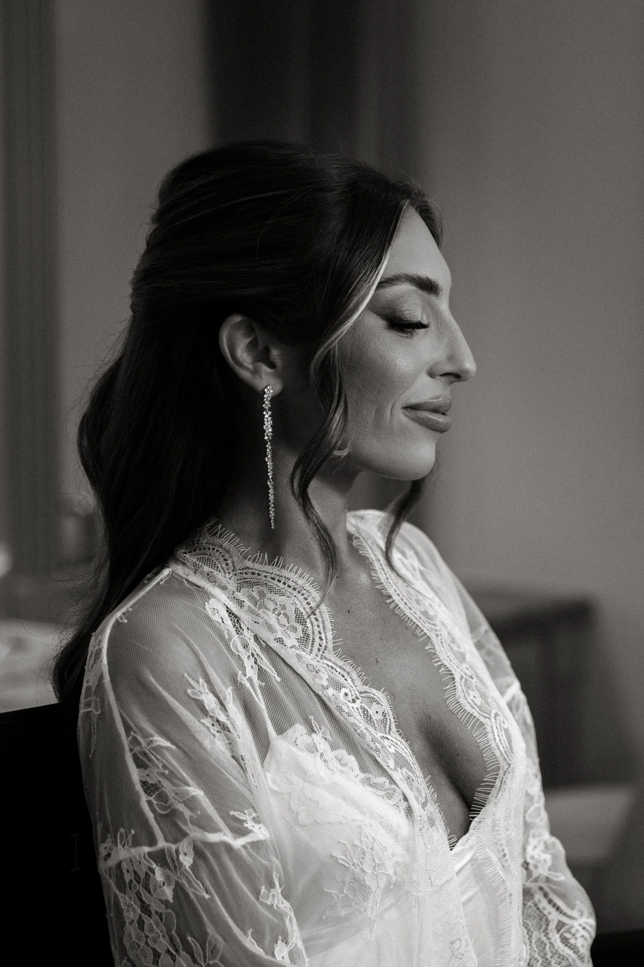 Black and white photo of a woman with long, wavy hair and closed eyes, wearing drop earrings and a lace dress with floral embroidery, captured from the side with a soft expression.