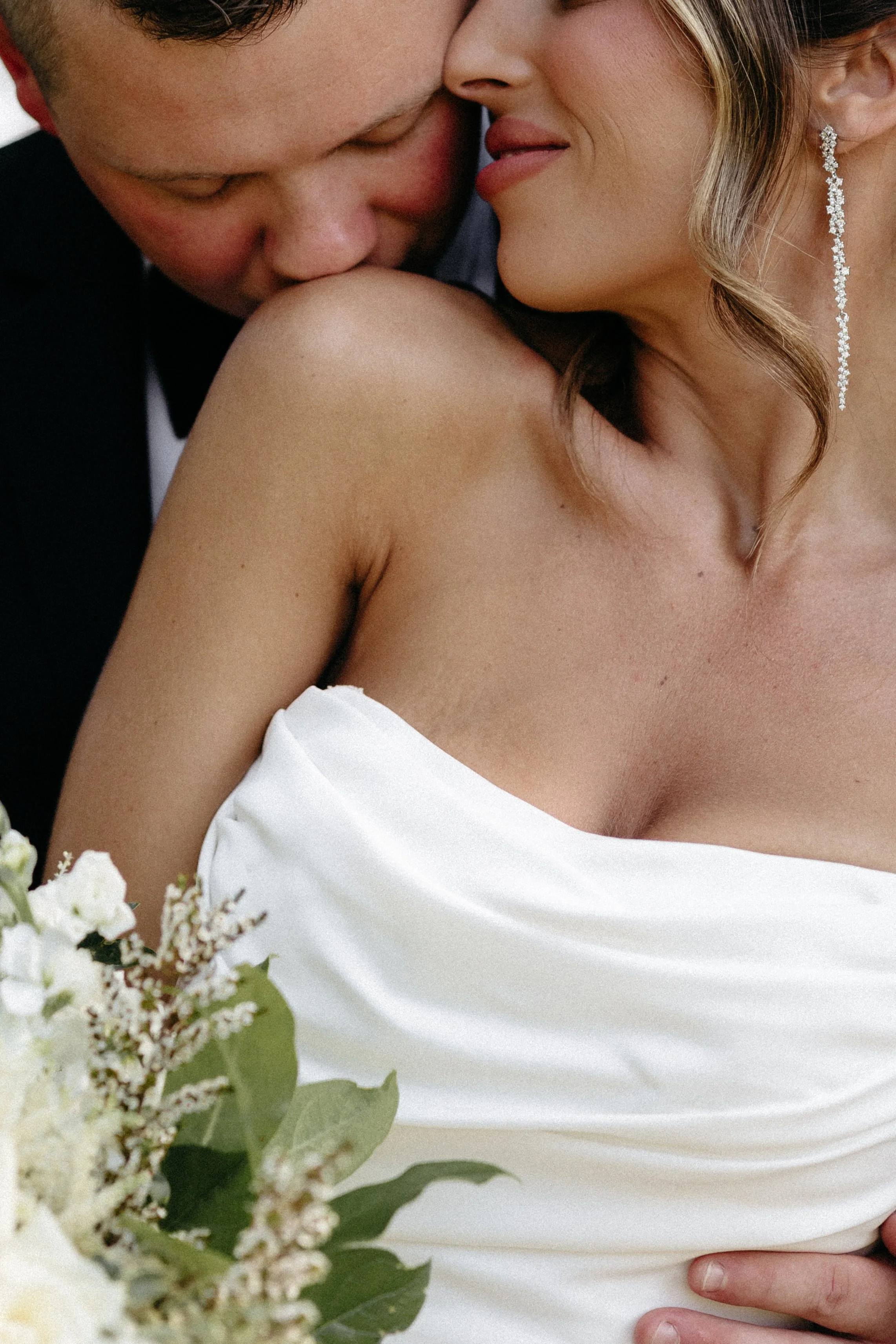 Close-up of a bride and groom embracing on their wedding day, with the bride wearing a white strapless wedding dress and dangling earrings, and the groom in a black tuxedo, near a bouquet of white flowers.