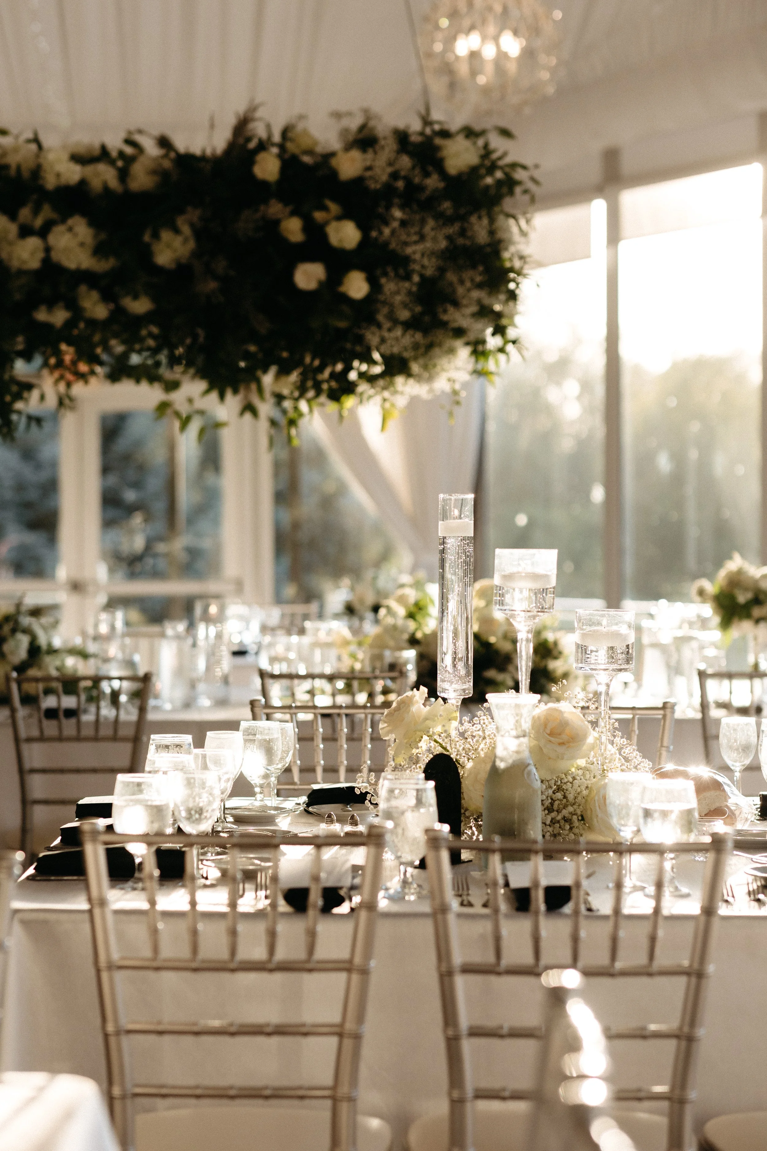 Elegant wedding reception table with white floral arrangements, glassware, and candles, illuminated by natural light.