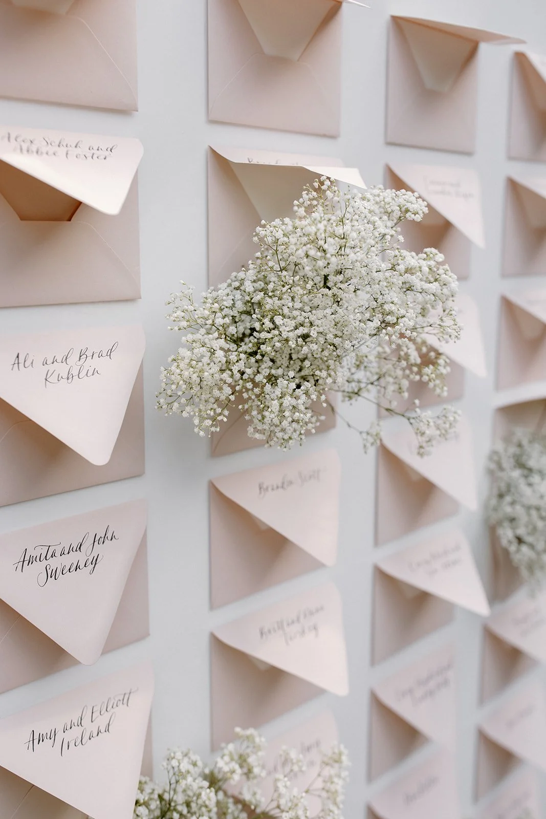 A wall with multiple pink envelope-style cards held open, each containing a folded card and handwritten names, with one card holding a bouquet of white baby's breath flowers.