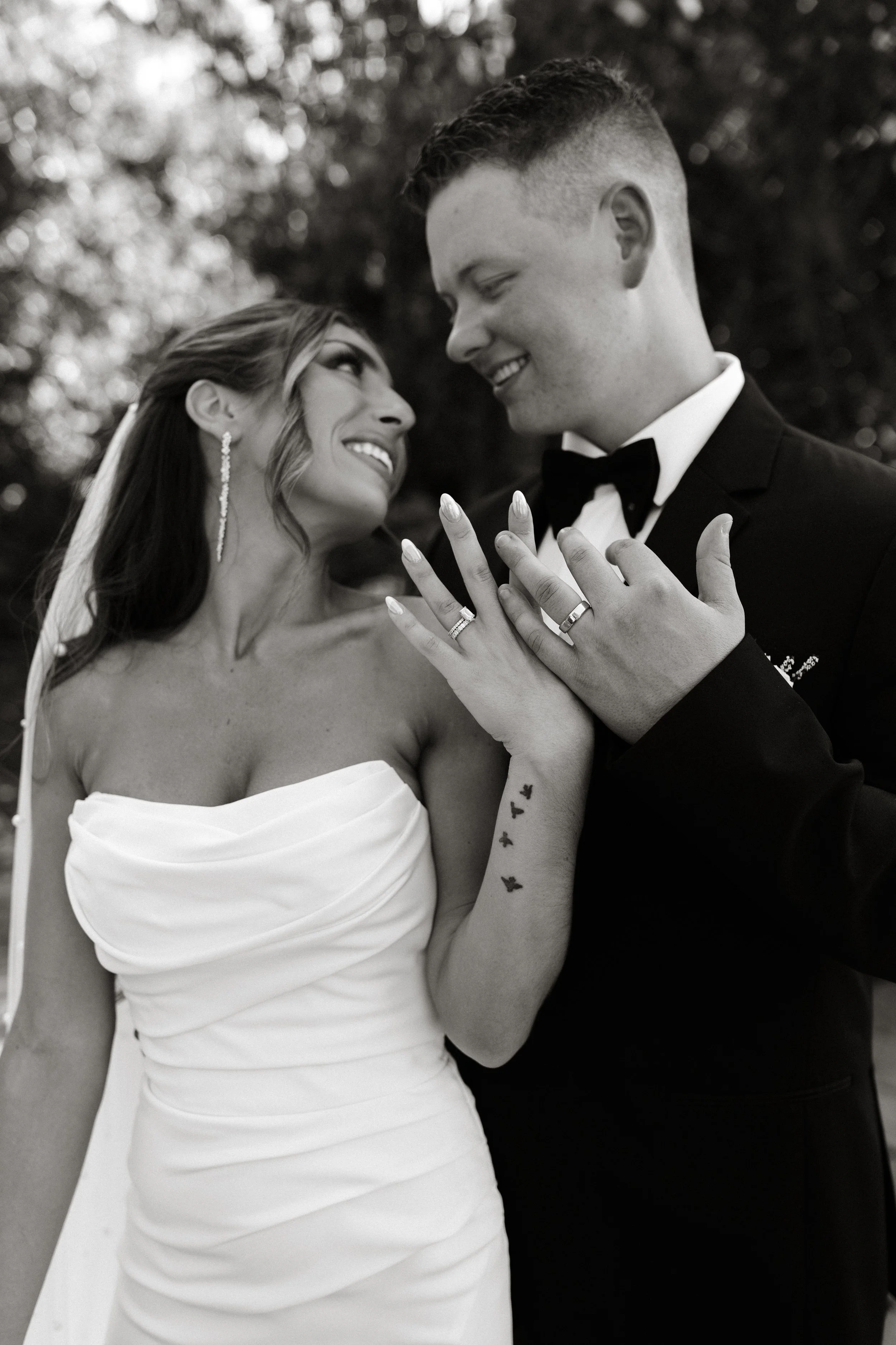 Black and white photo of a bride and groom looking at each other and smiling, with their hands together showing wedding rings, outdoors with blurred trees in the background.