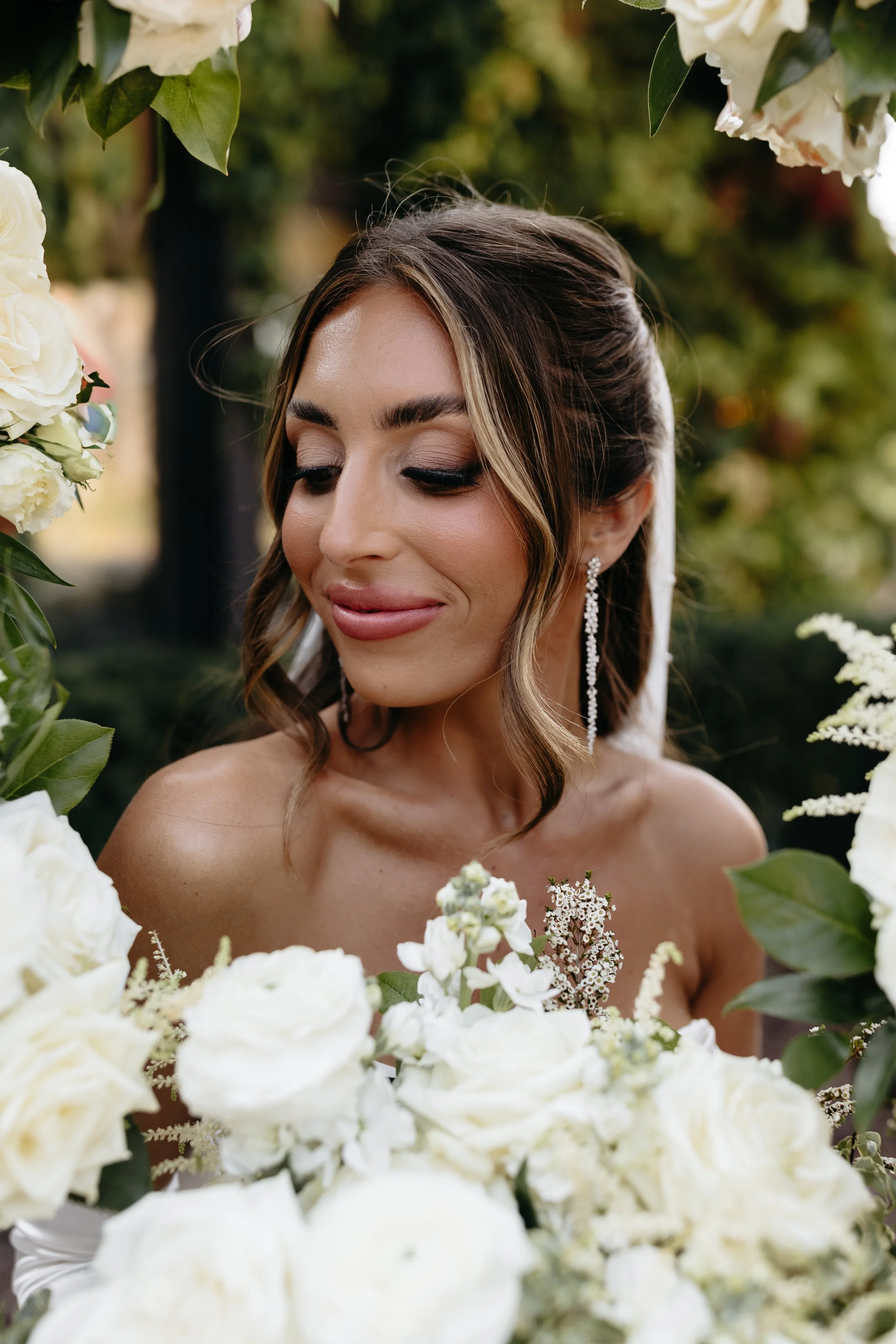 A woman with makeup and jewelry surrounded by white flowers outdoors.