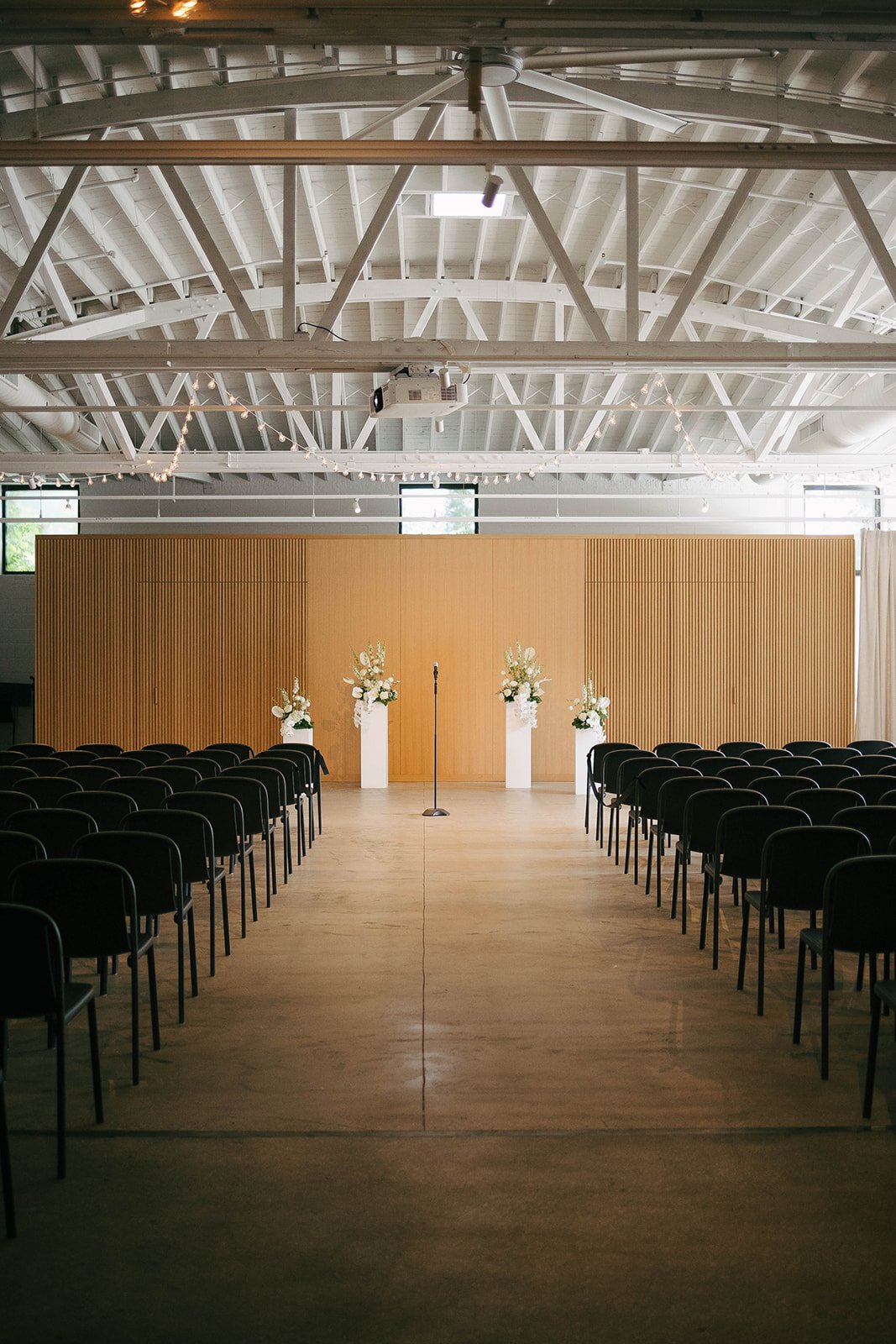 An indoor wedding ceremony setup with black chairs arranged in rows facing a wooden backdrop, decorated with white flowers on tall white vases, a microphone stand in the center, and string lights hanging from the ceiling.