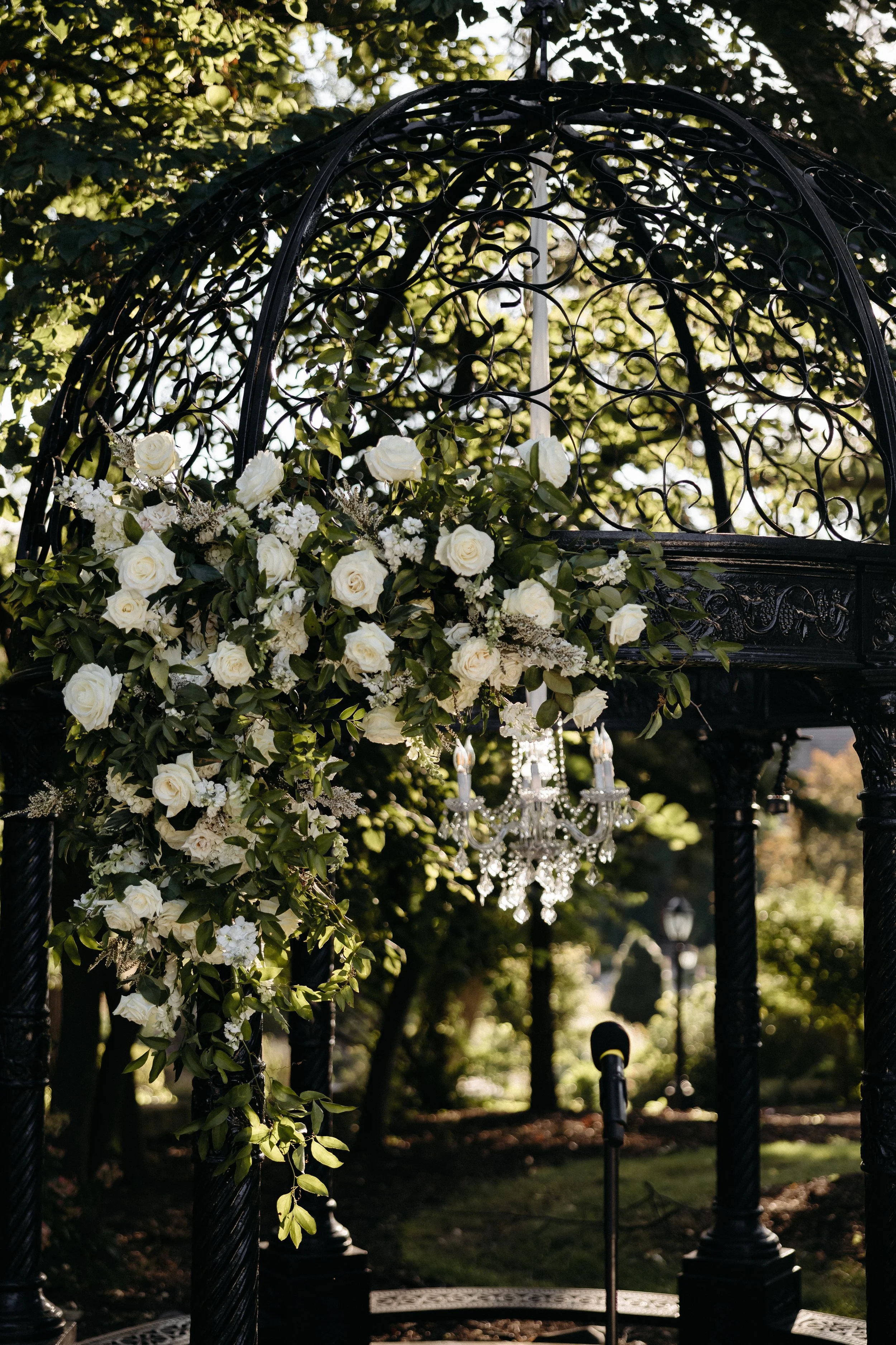 A decorative Black metal garden arch decorated with white roses, greenery, and a chandelier hanging inside in an outdoor garden setting.