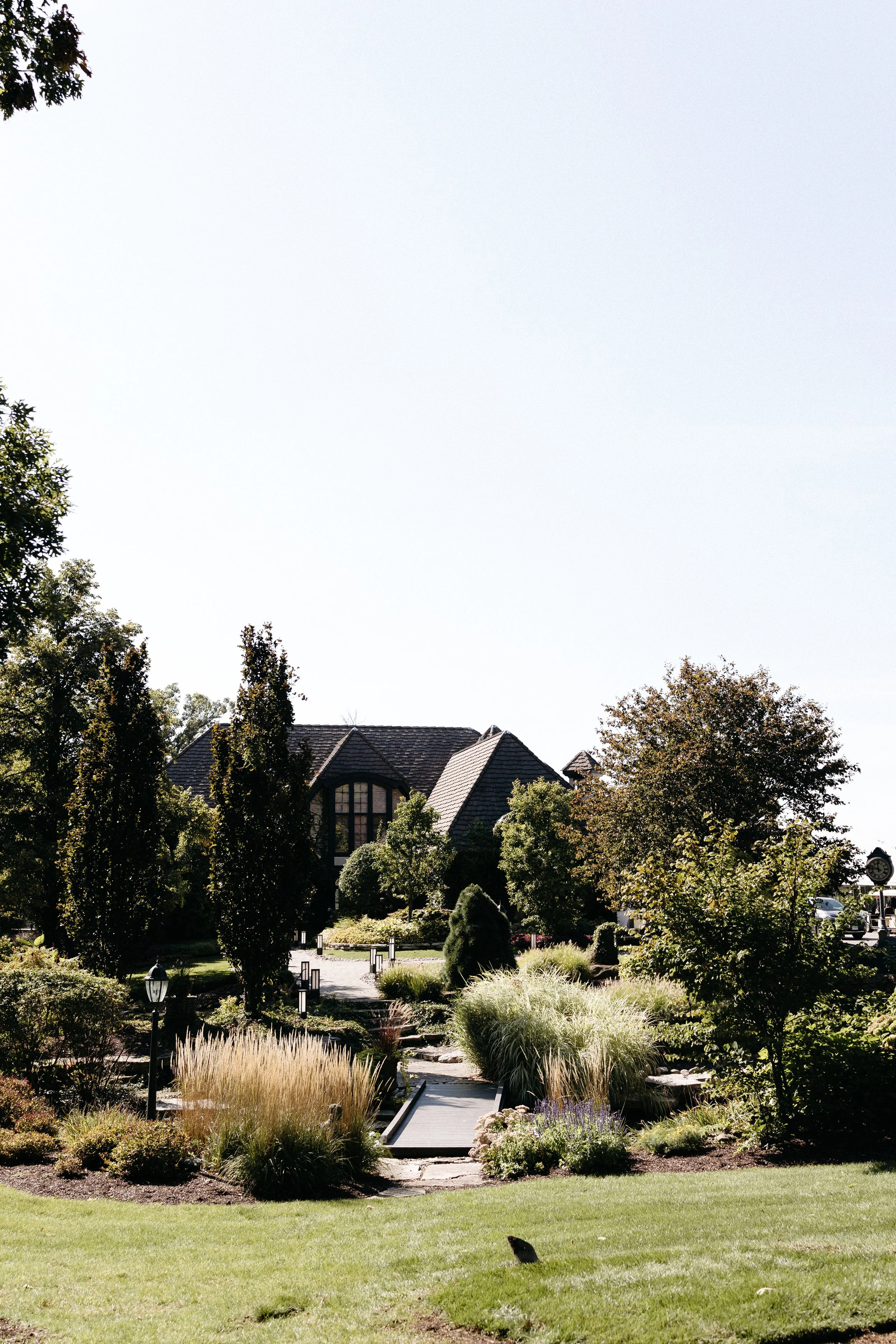 A landscaped garden with a variety of shrubs, trees, and ornamental grasses, leading up to a large house with dark roofing and arched windows, under a clear sky.
