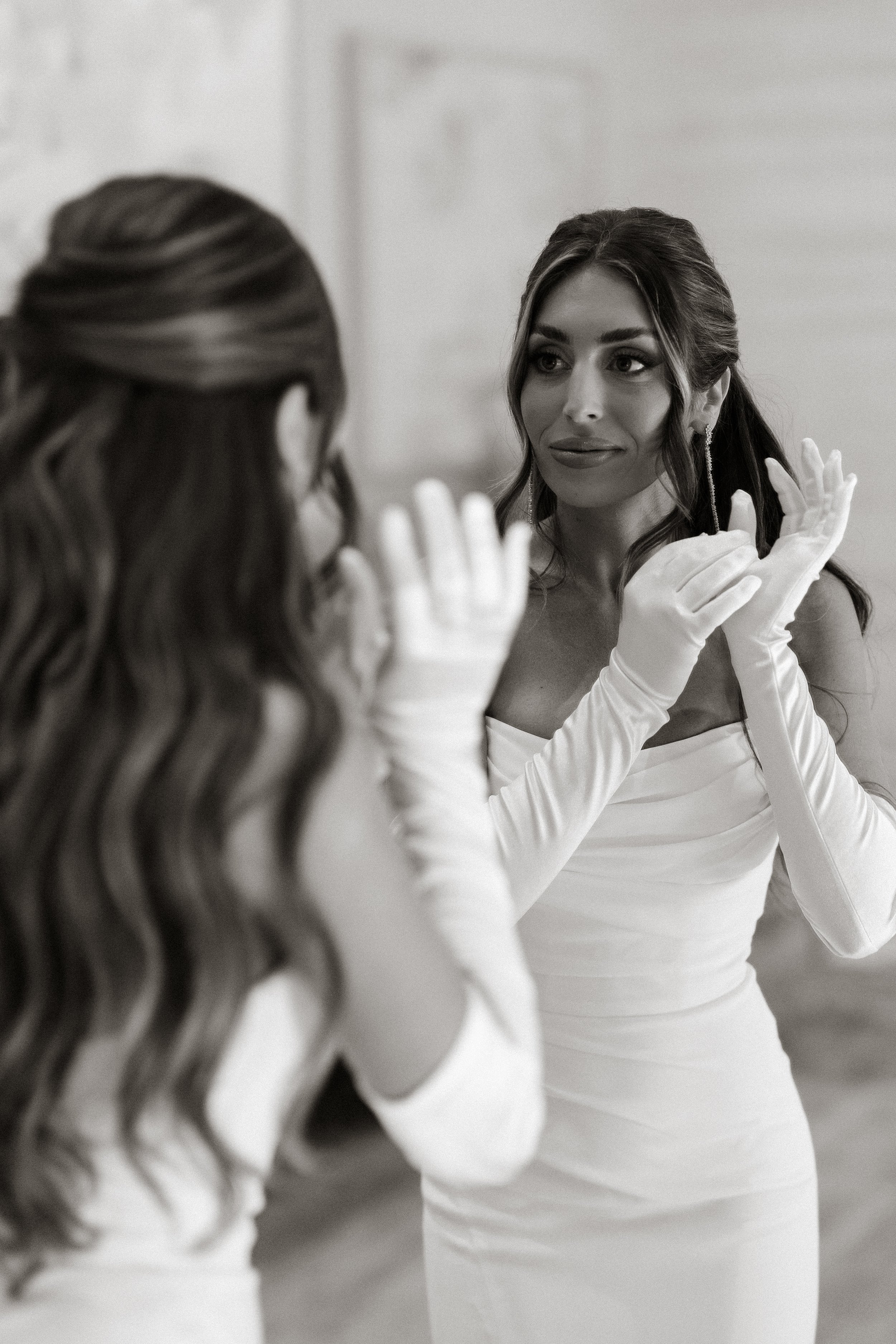Black and white photograph of a woman in a white dress adjusting her gloves while another woman in a white dress looks on, both in an indoor setting.