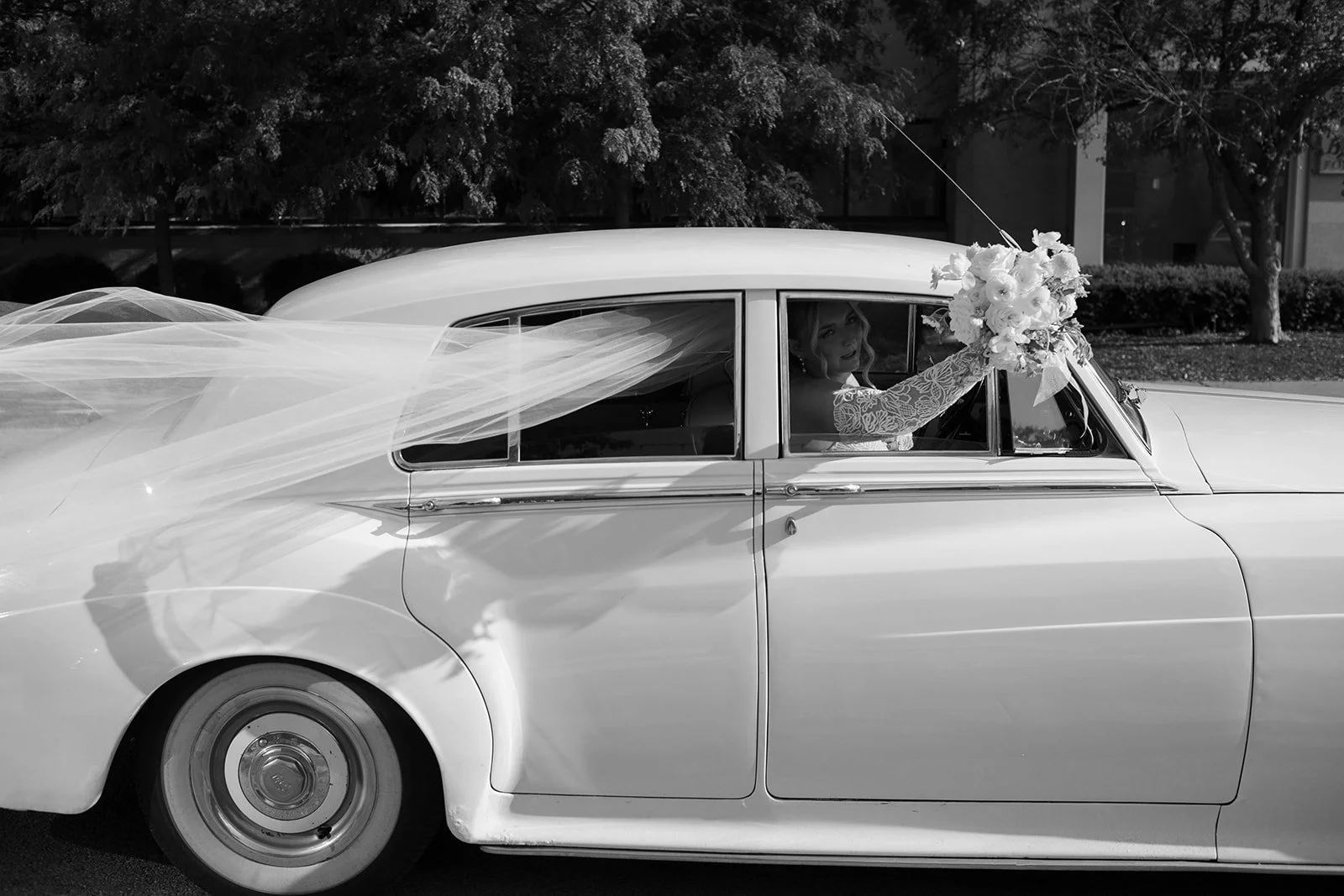 A black and white photo of a bride sitting in the backseat of a vintage white car, holding a bouquet of flowers out of the window. The car has a flowing veil or fabric attached to it, and there are trees and bushes in the background.