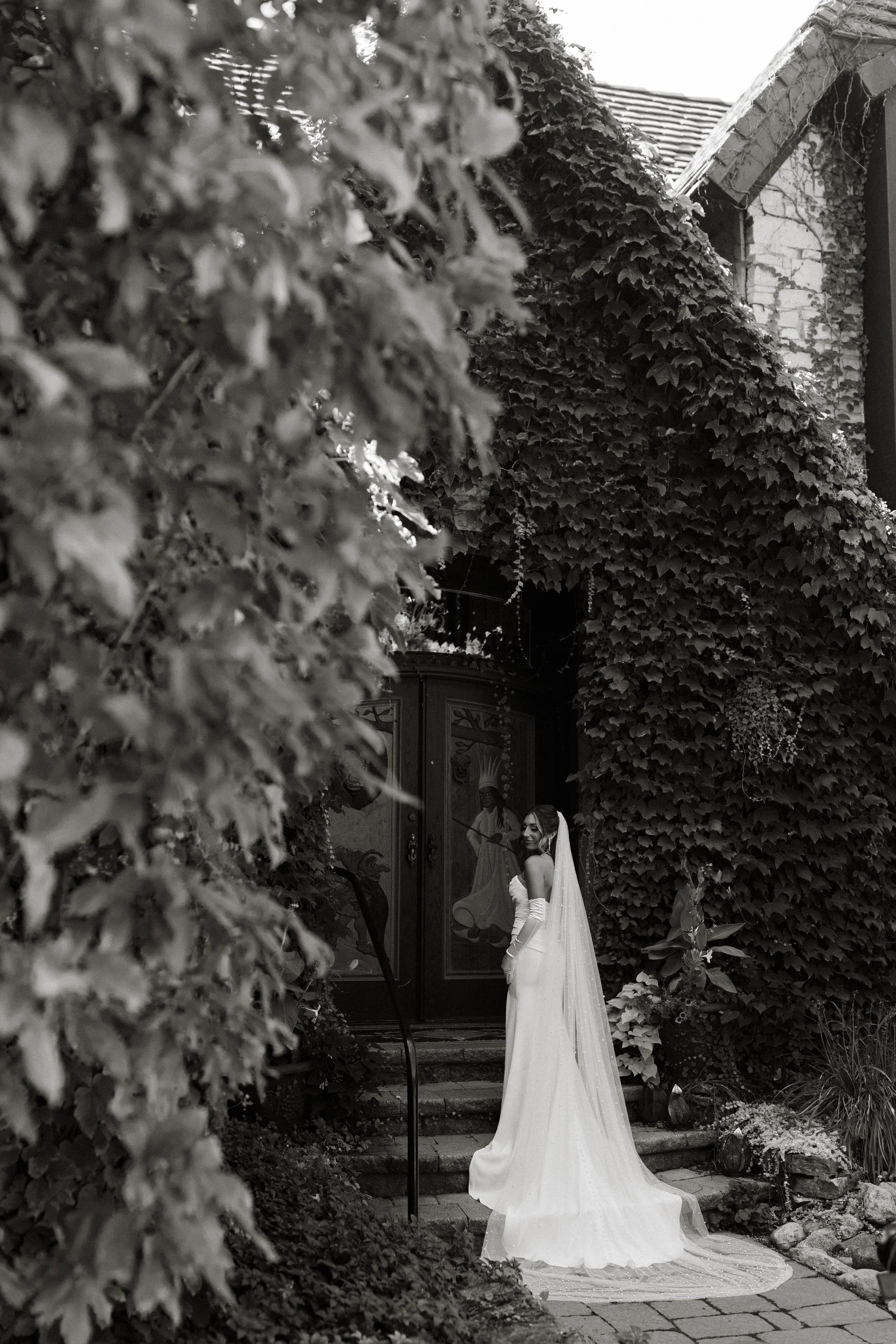 A bride in a white wedding dress with long gloves and veil standing on steps outside a building covered in ivy, with a mural of a woman playing a stringed instrument painted on the door.