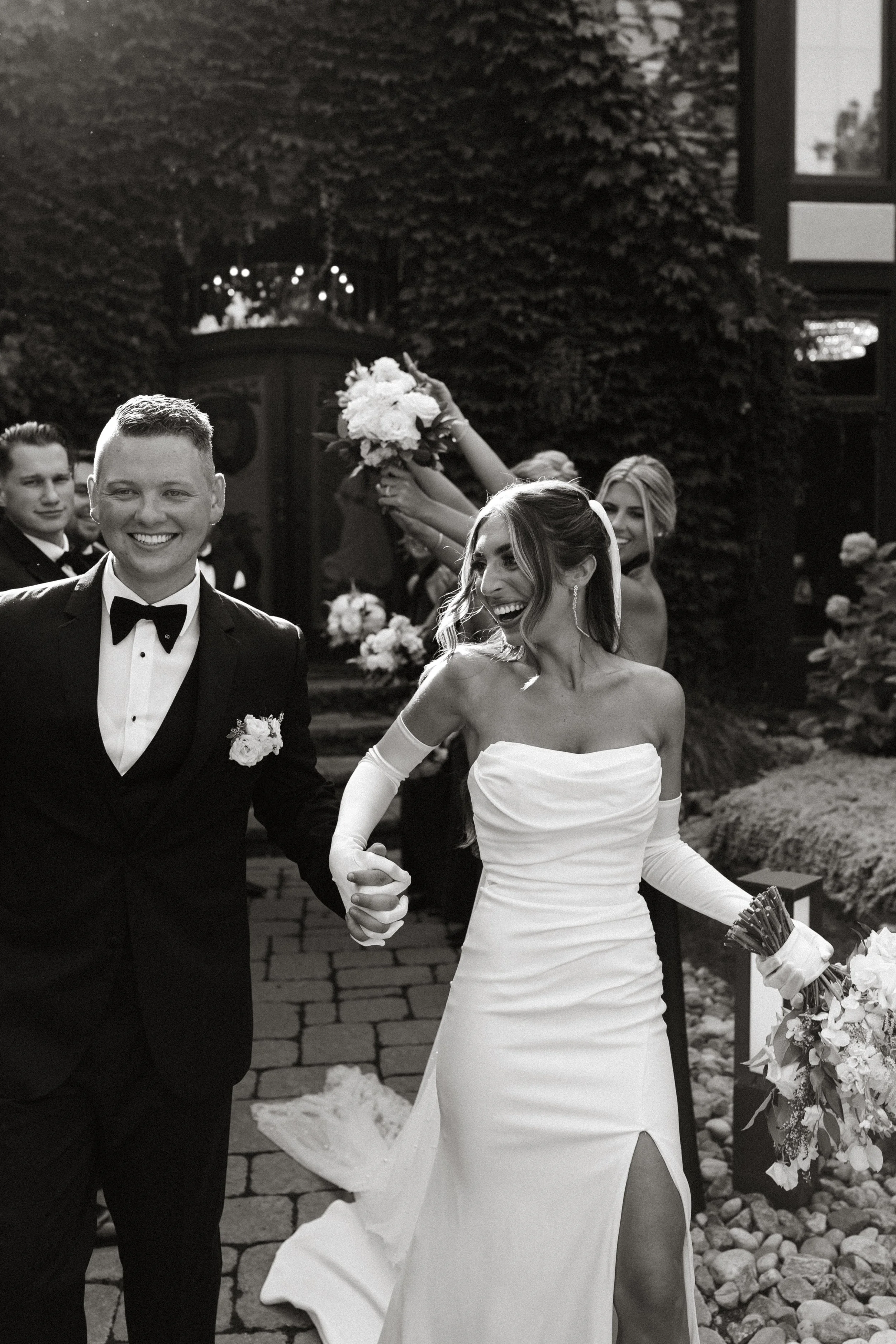 Black and white photo of a wedding celebration showing a smiling bride and groom holding hands, with friends or family in the background. The bride wearing a strapless wedding gown and gloves, and the groom in a tuxedo with a bowtie, celebrating joyf
