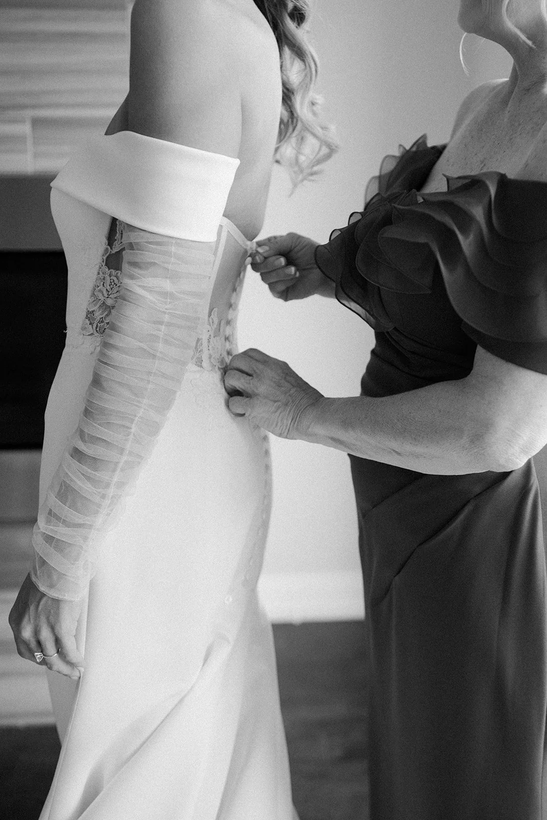 A woman in a wedding dress has her corset being fastened by another woman, possibly her mother, in an indoor setting.