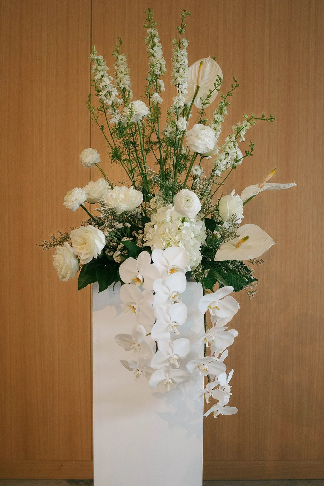 A tall white floral arrangement in a white vase with white roses, orchids, calla lilies, and other white flowers, set against a wooden background.