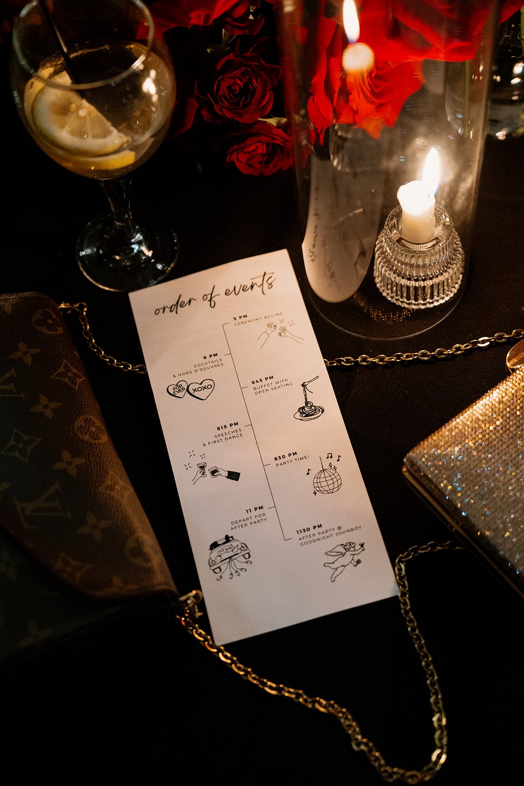 A wedding or party event setup featuring a glass of lemon water, a bouquet of red roses, a lit candle in a glass holder, a gold designer clutch, a gold chain, and a printed schedule of events.