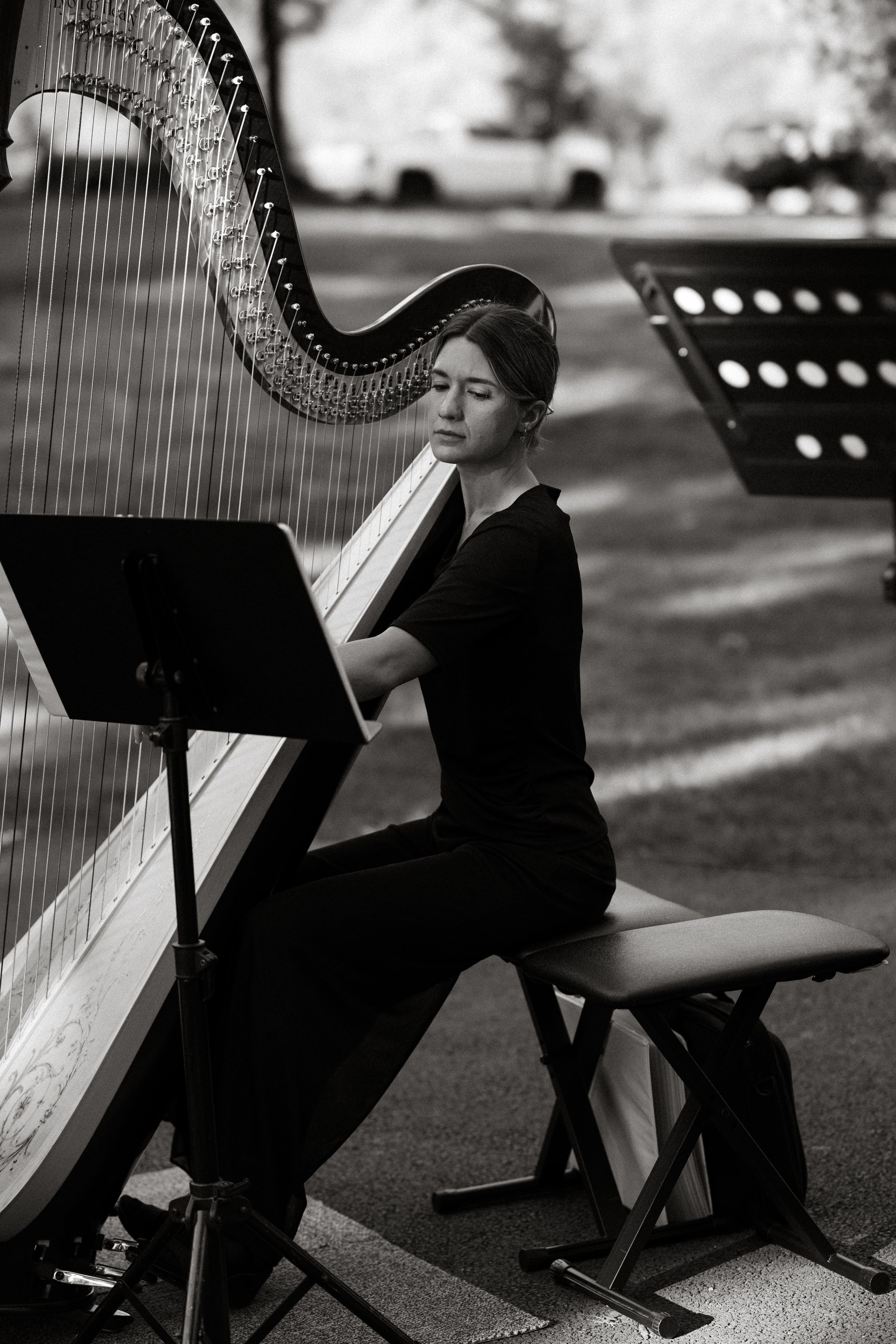A woman playing a large pedal harp outdoors, sitting on a bench with music stand in front of her, with a blurred background of trees and a parking lot.
