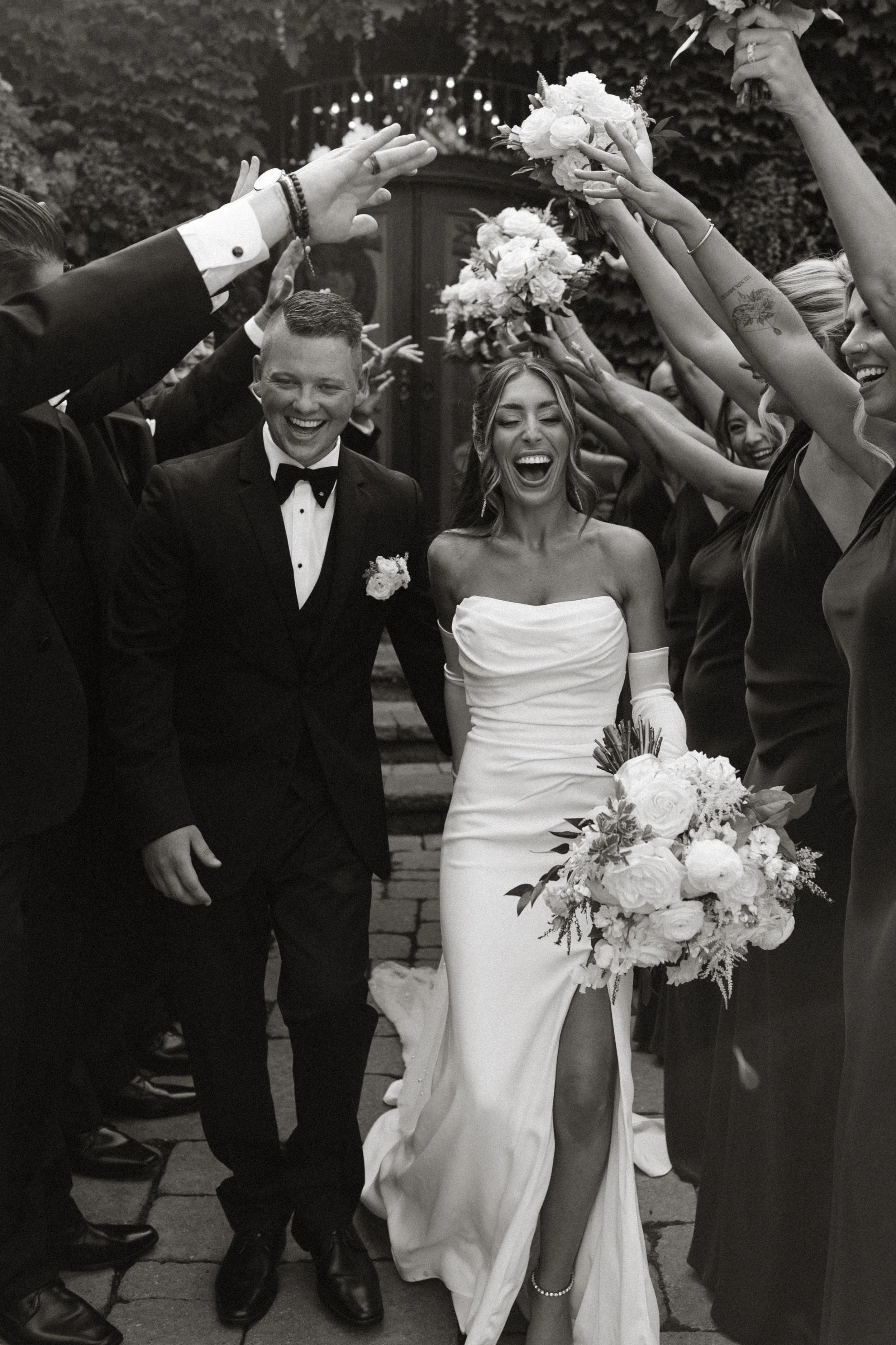 A bride in a strapless wedding gown holding a bouquet, walking beside a groom in a tuxedo, surrounded by friends in a celebratory outdoor wedding procession with raised bouquets.