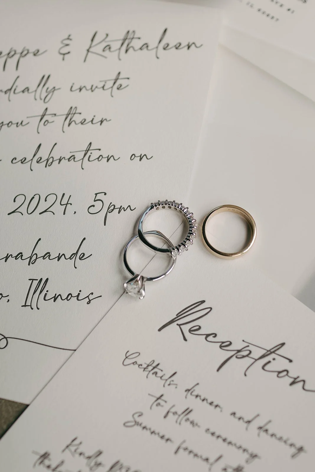 Invitation card with elegant handwritten text, wedding rings placed on top, one with a gemstone and another plain.