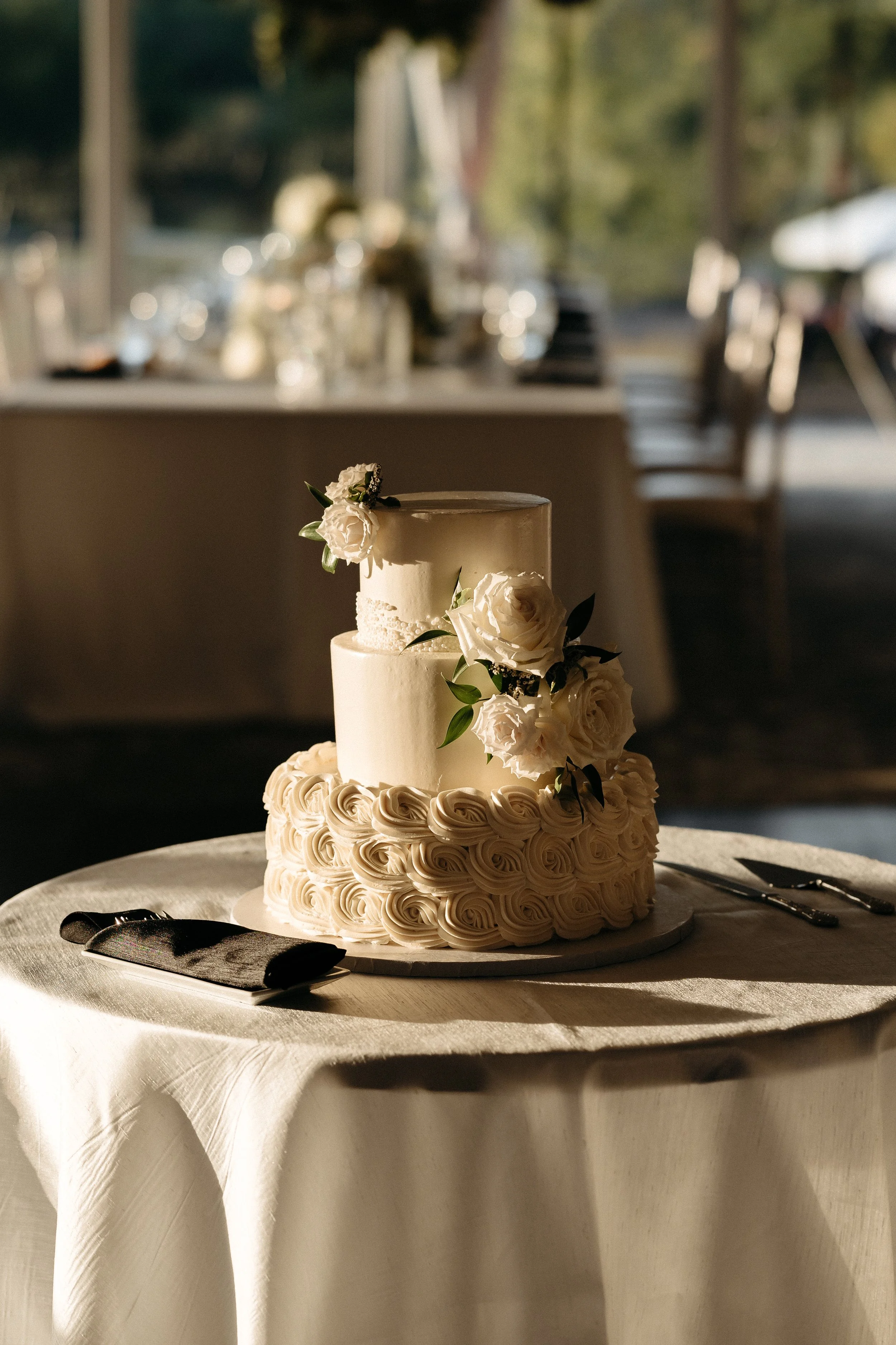 A three-tier white wedding cake decorated with white roses and green leaves, placed on a round table with a white tablecloth at a wedding reception.