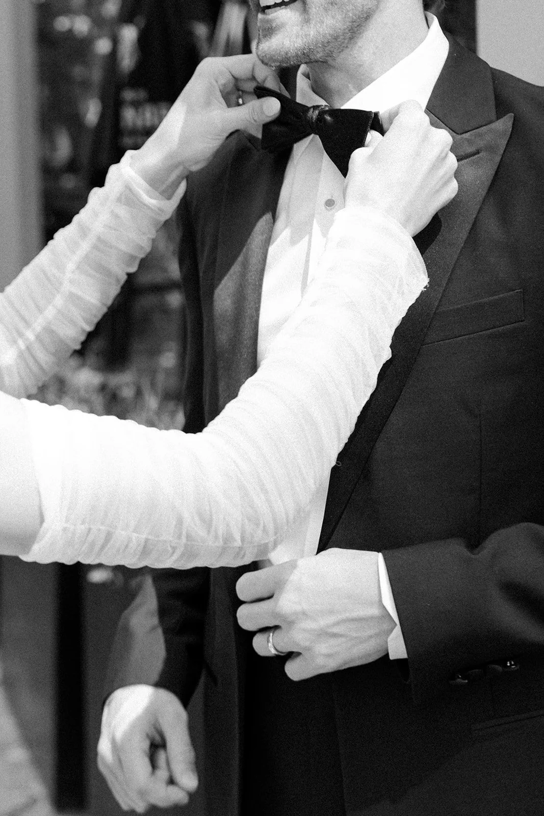 A person helping a man adjust his black bow tie while he wears a tuxedo. The person assisting wears a long-sleeved shirt, and the man in tuxedo is adjusting the tie in front of a blurred background.