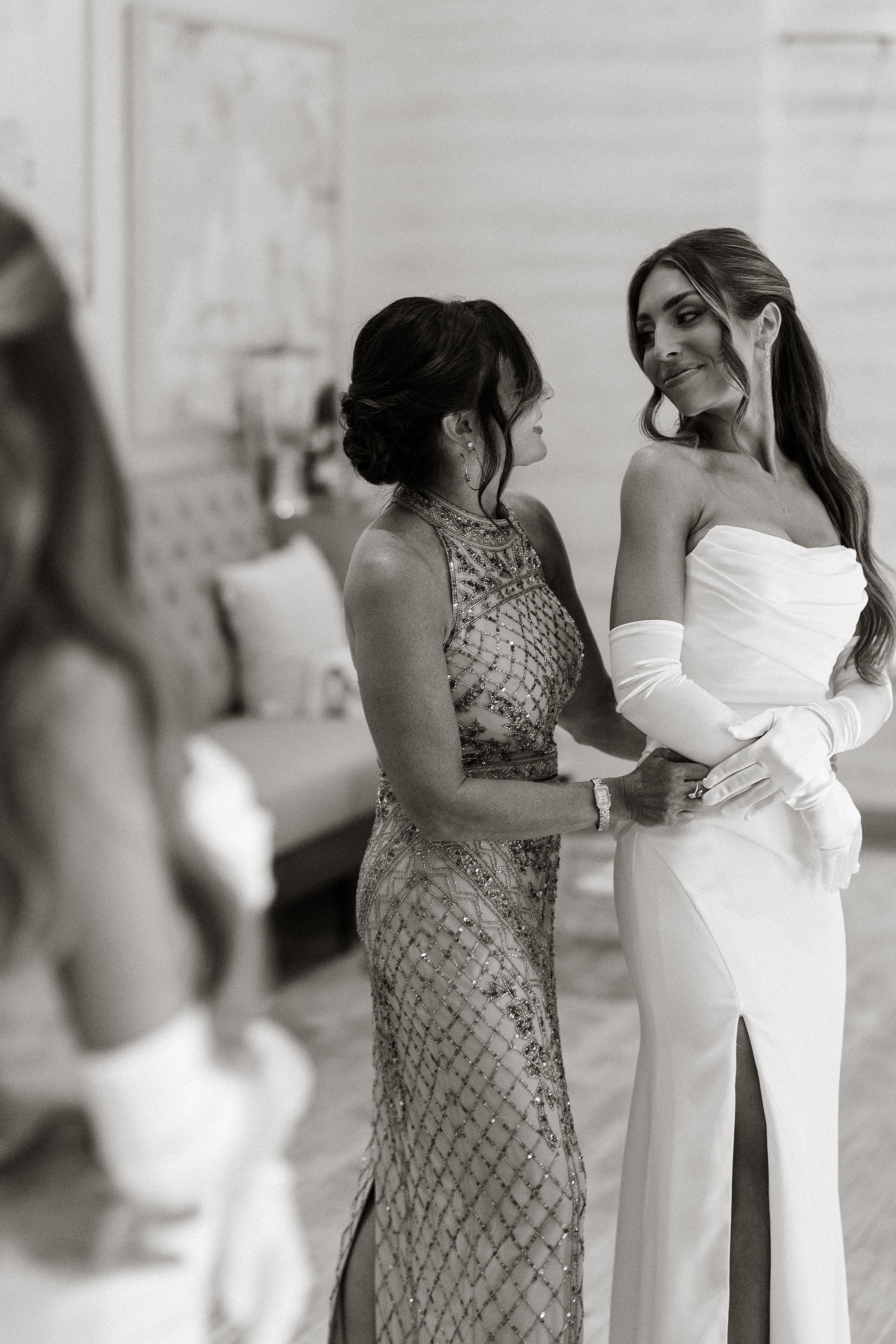 A woman in a white wedding dress with long gloves stands with an older woman in an ornate dress, holding her hand, at a wedding ceremony.