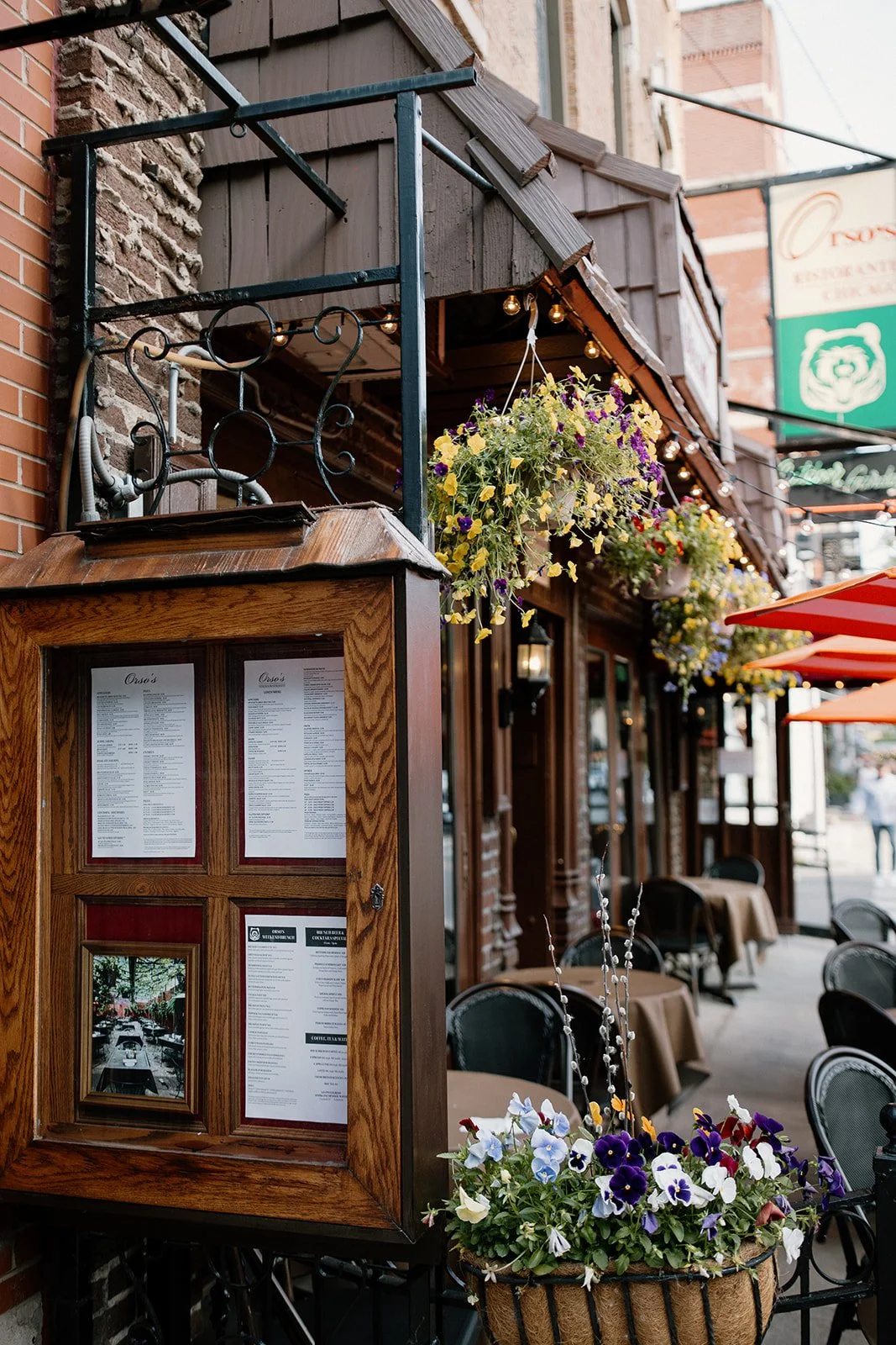 Outdoor restaurant with hanging flower baskets, a wooden menu display case, black metal chairs, and tables with brown tablecloths under red umbrellas.
