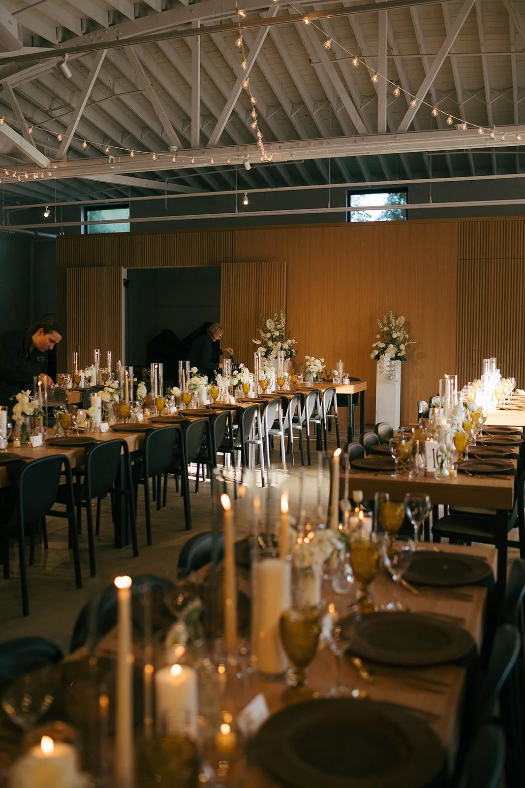 Elegant banquet setup with long tables decorated with candles, flowers, and glassware, ready for an event in a modern venue with string lights on the ceiling.