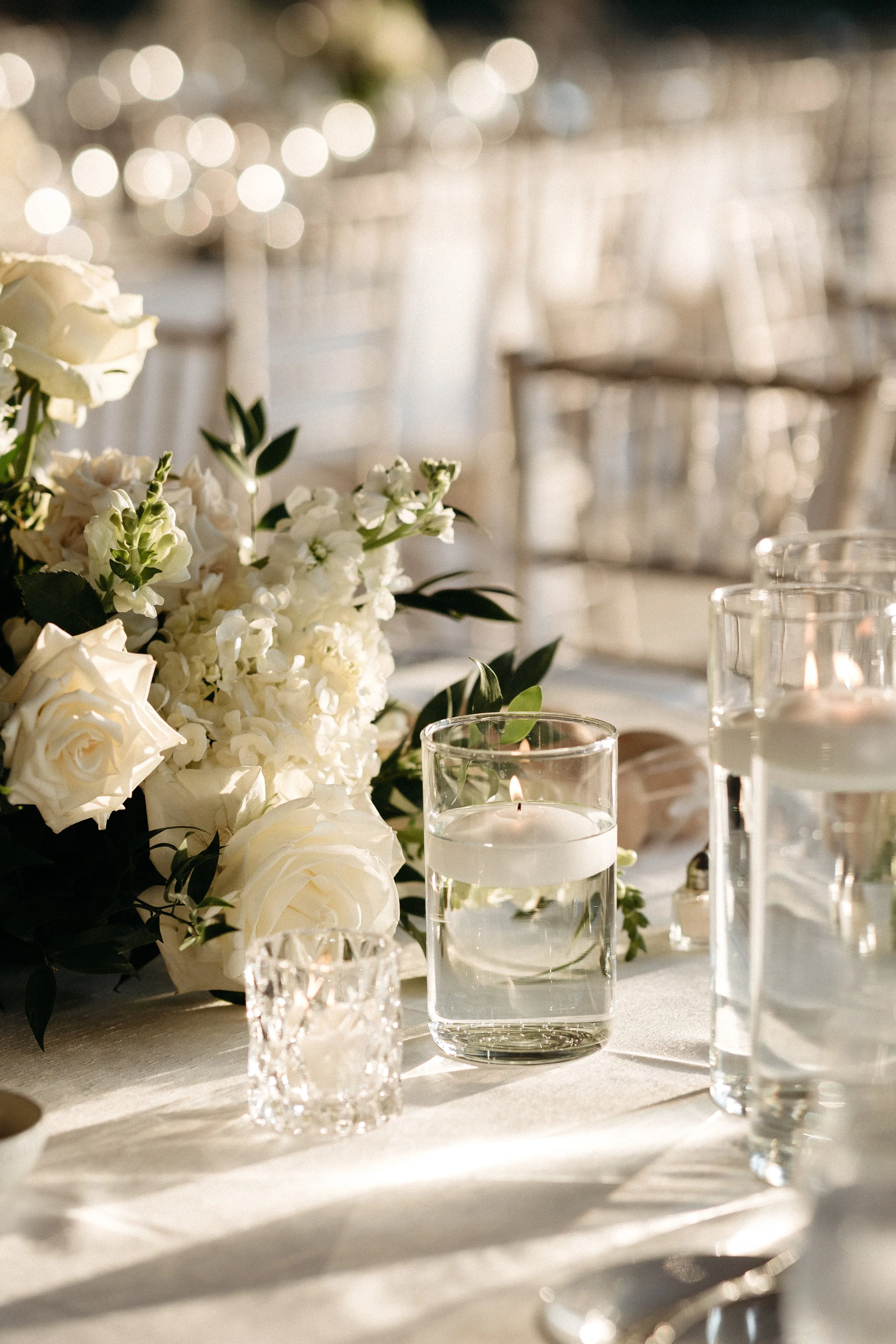 Decorative table setting with white flowers, candles in glass holders, and clear glasses on a table at a bright, elegant event.