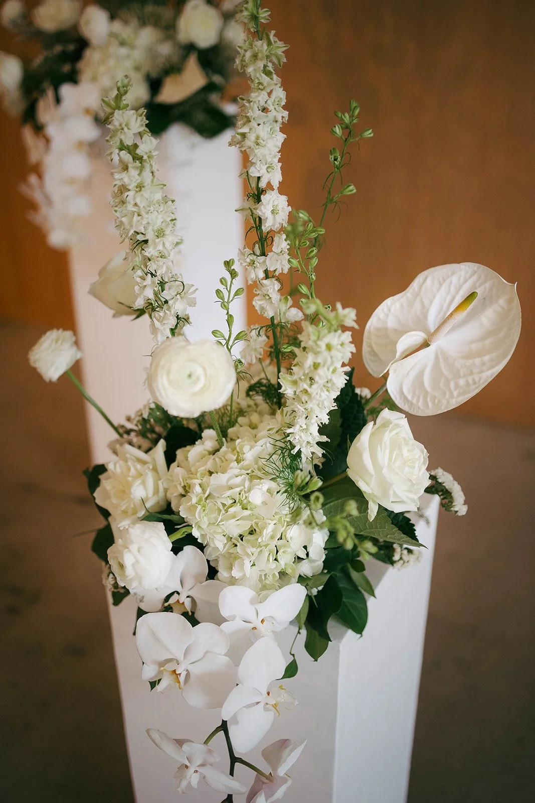 A tall white flower arrangement with white roses, orchids, hydrangeas, and greenery in a white vase.