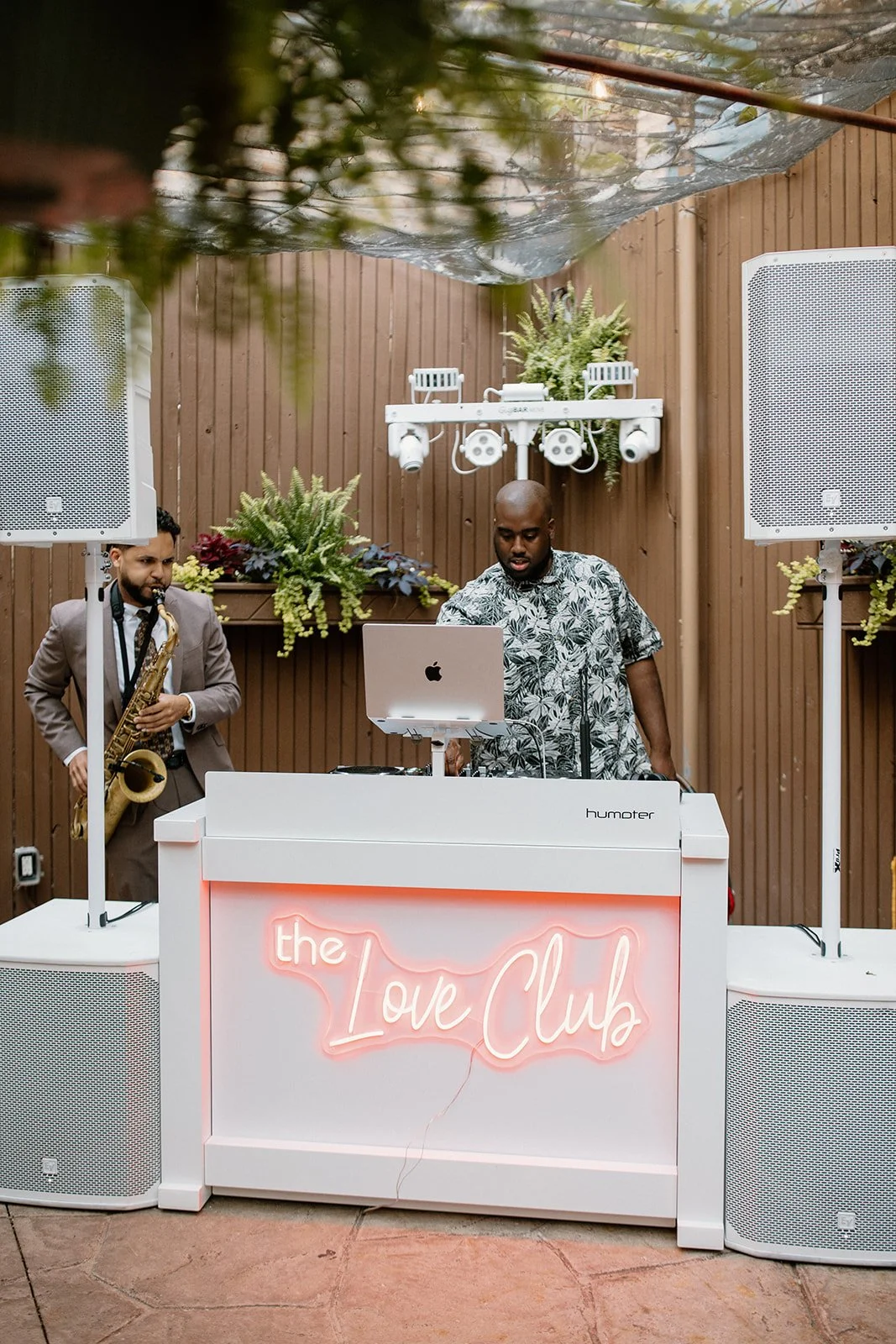 Two musicians, one playing a saxophone and the other operating a DJ setup, performing at a venue called the Love Club, with wooden wall backdrop, decorative greenery, and a neon sign.