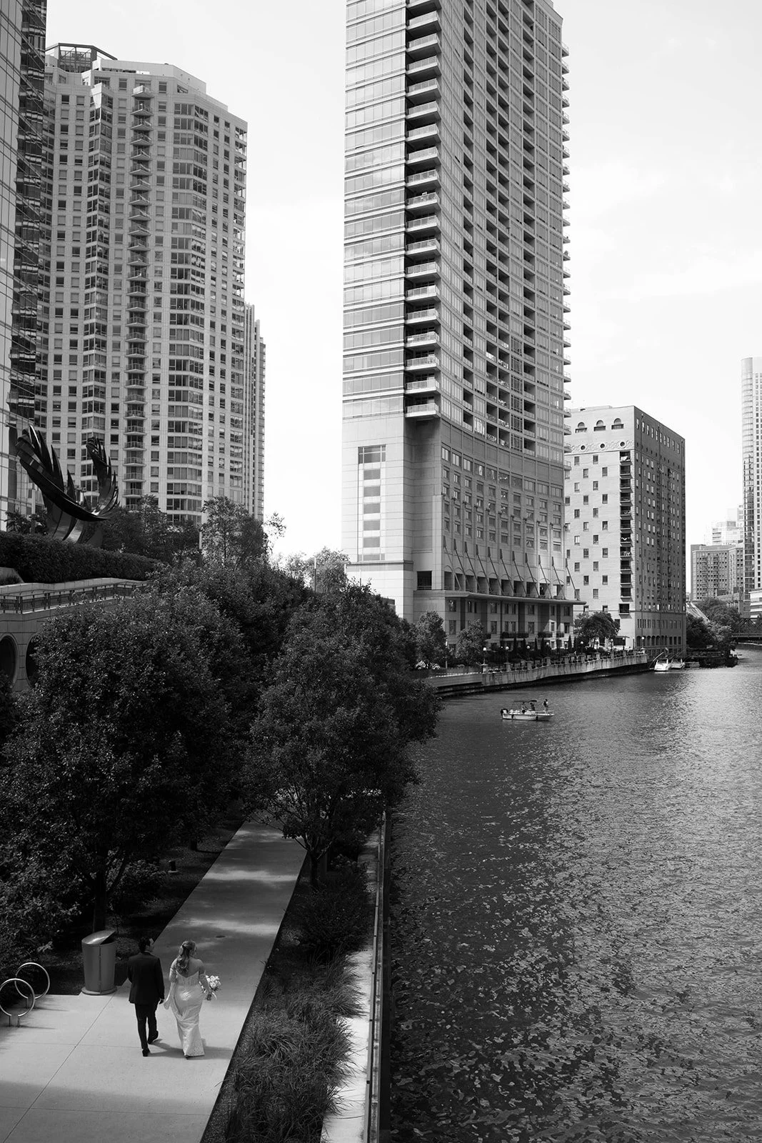 A black-and-white photo of a city waterfront with high-rise buildings, trees, and a walkway along the water where a couple is walking, with a boat on the river.