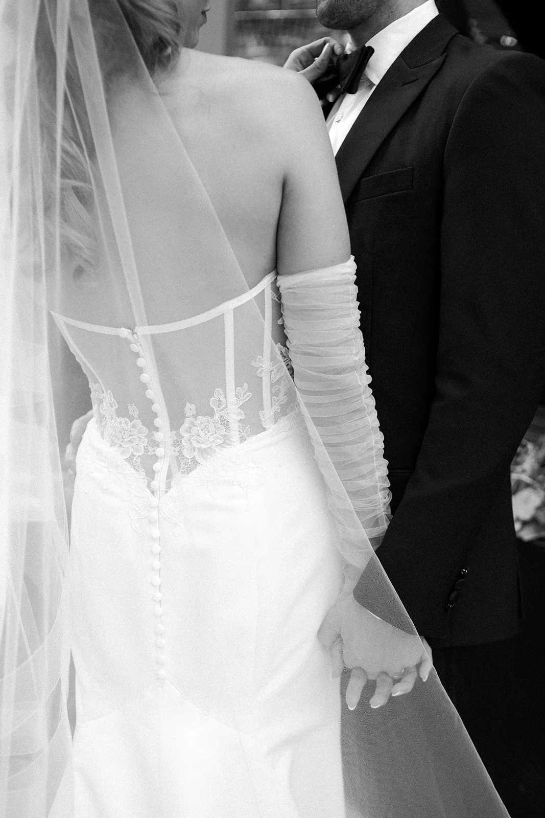 Close-up of a bride and groom holding hands during a wedding ceremony, with the bride wearing a lace wedding dress and the groom in a black tuxedo.
