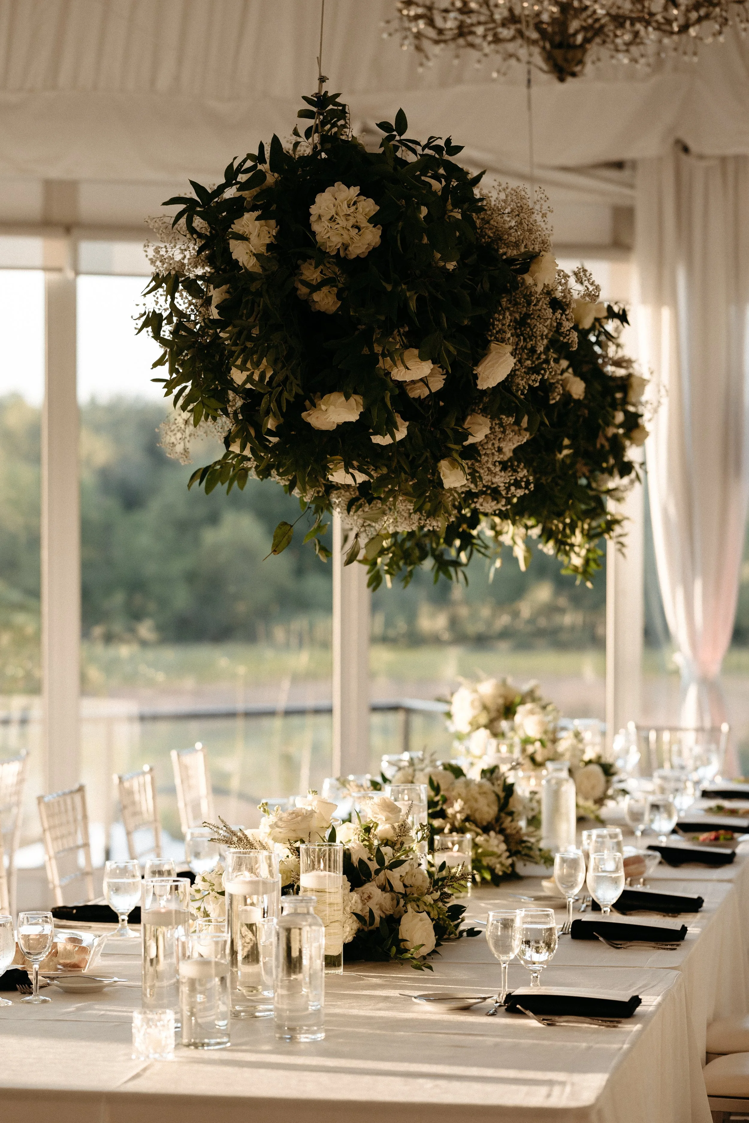 Elegant wedding reception table with white floral centerpieces and tall hanging flower arrangement in a bright room with large windows and white curtains.