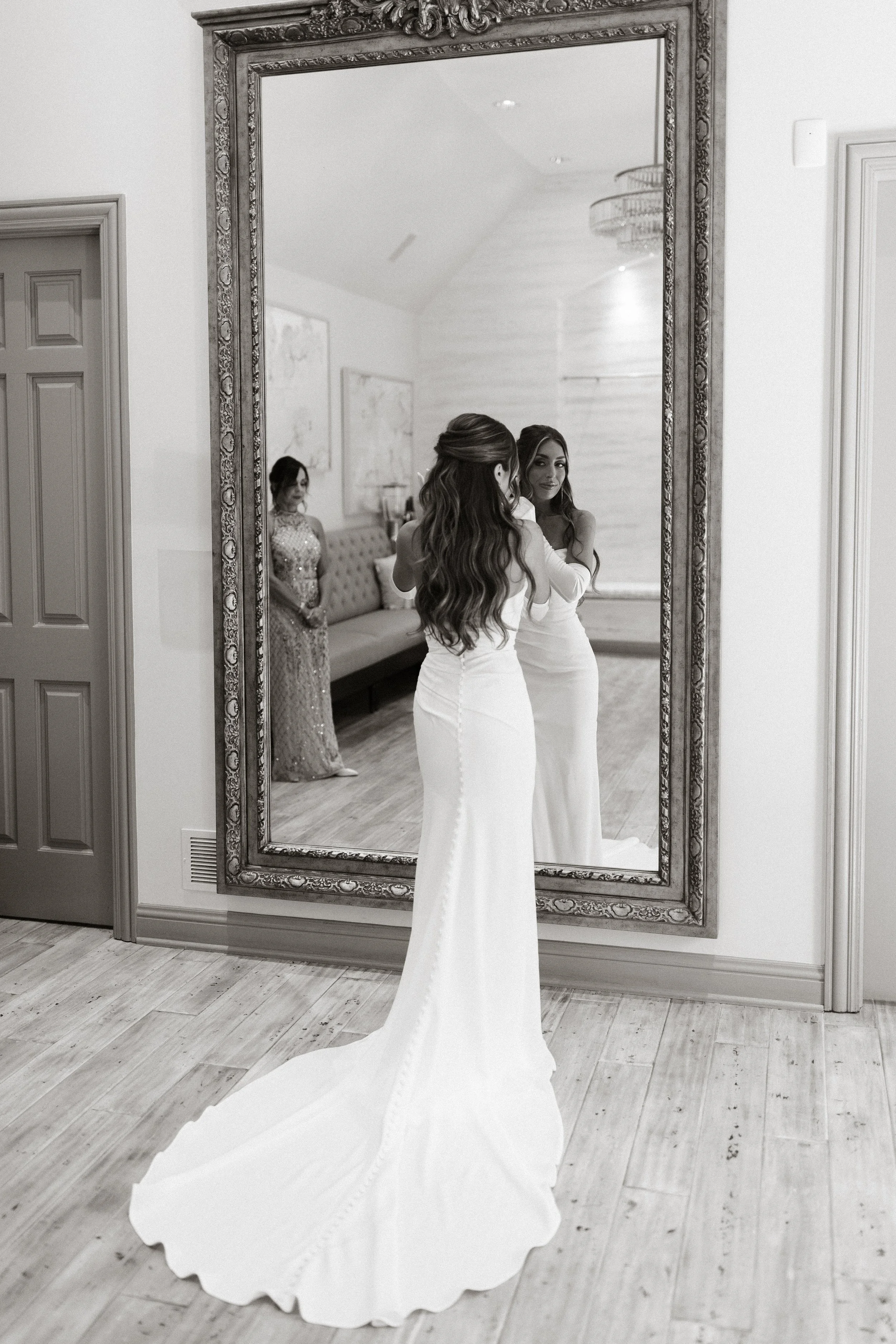 Two women in wedding dresses looking at themselves in a large ornate mirror, with a third woman in a decorative dress standing nearby in a room with light-colored walls and wood flooring.
