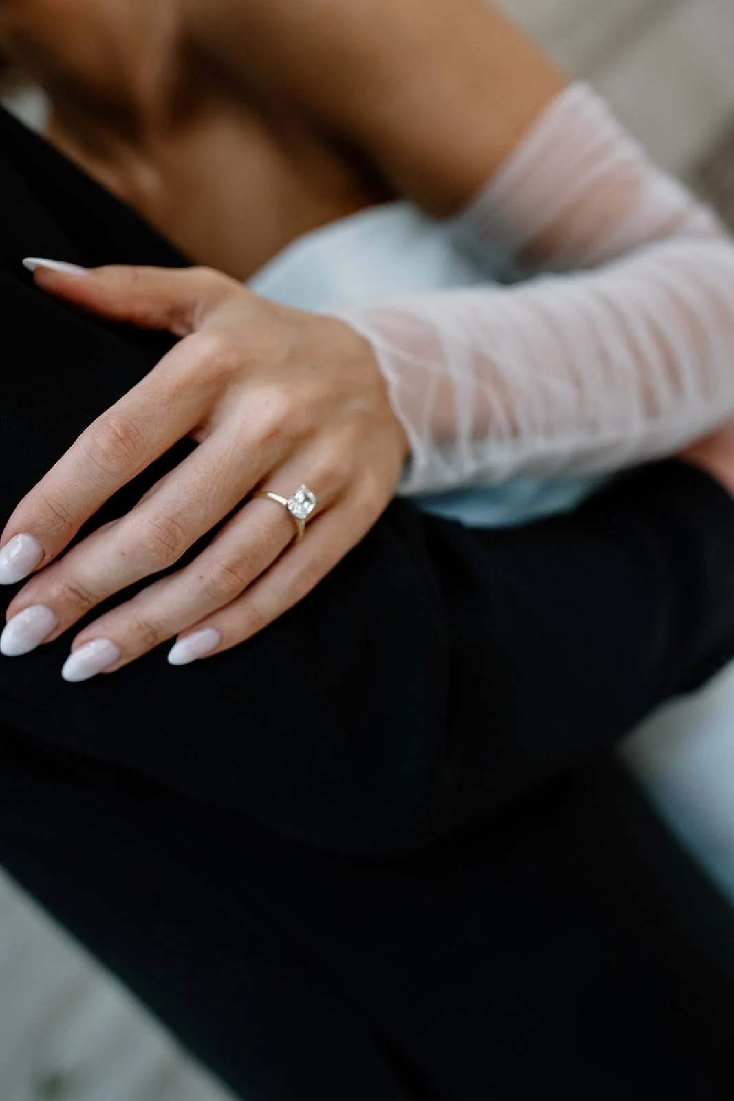 Close-up of a woman's hand with an engagement ring resting on a man's leg. The woman wears a sheer, long-sleeved white top, and her nails are painted in a light color.