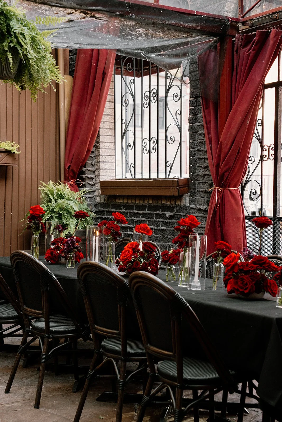 Dining table decorated with red flowers and candles, set in a brick-walled room with red curtains and a window with wrought iron bars.