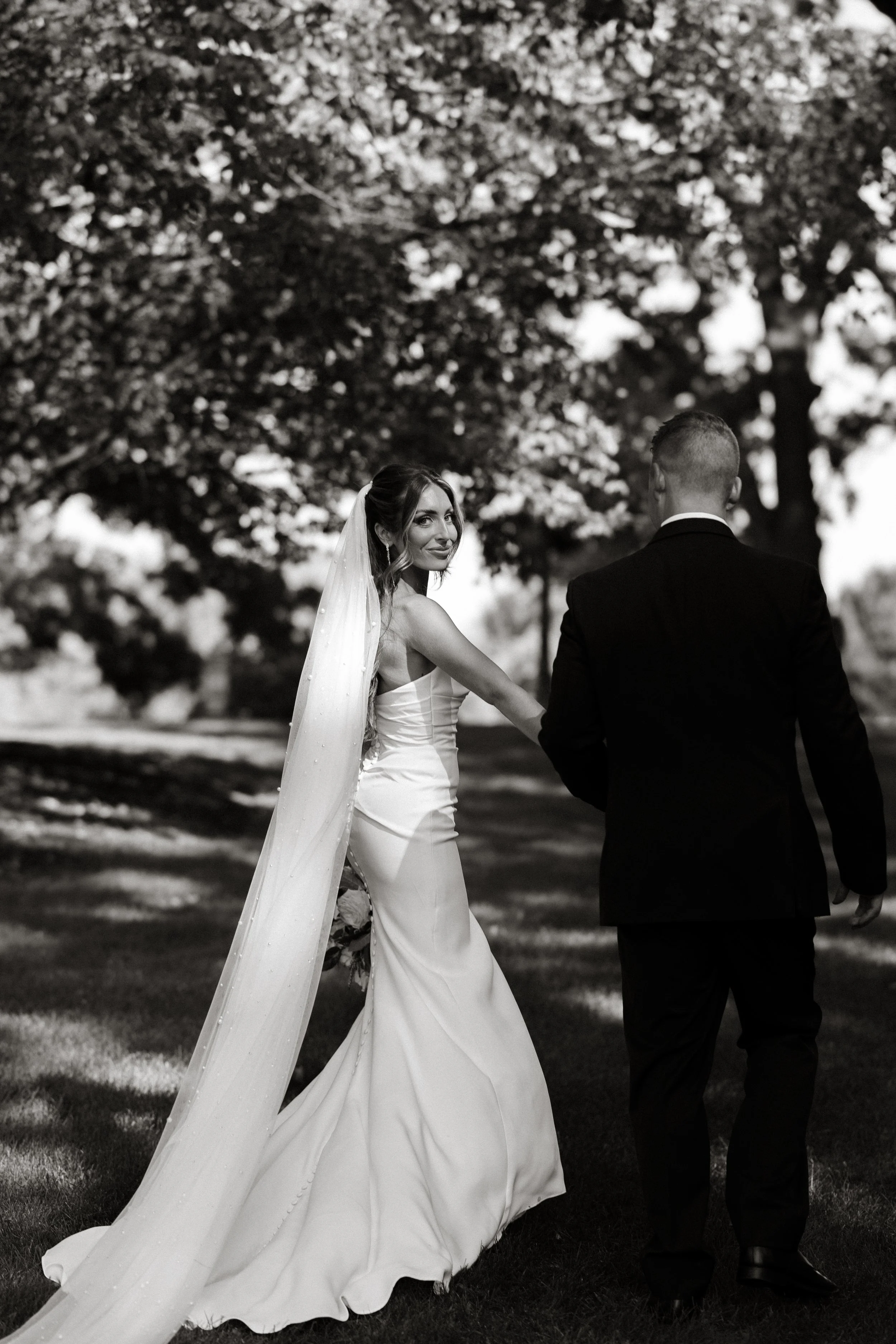 A black and white photo of a bride and groom holding hands outdoors with trees in the background.