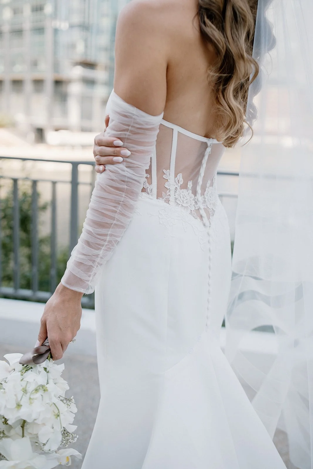 Bride in a white wedding gown with lace details on the back, holding a bouquet of white flowers, standing outdoors near a railing with buildings in the background.
