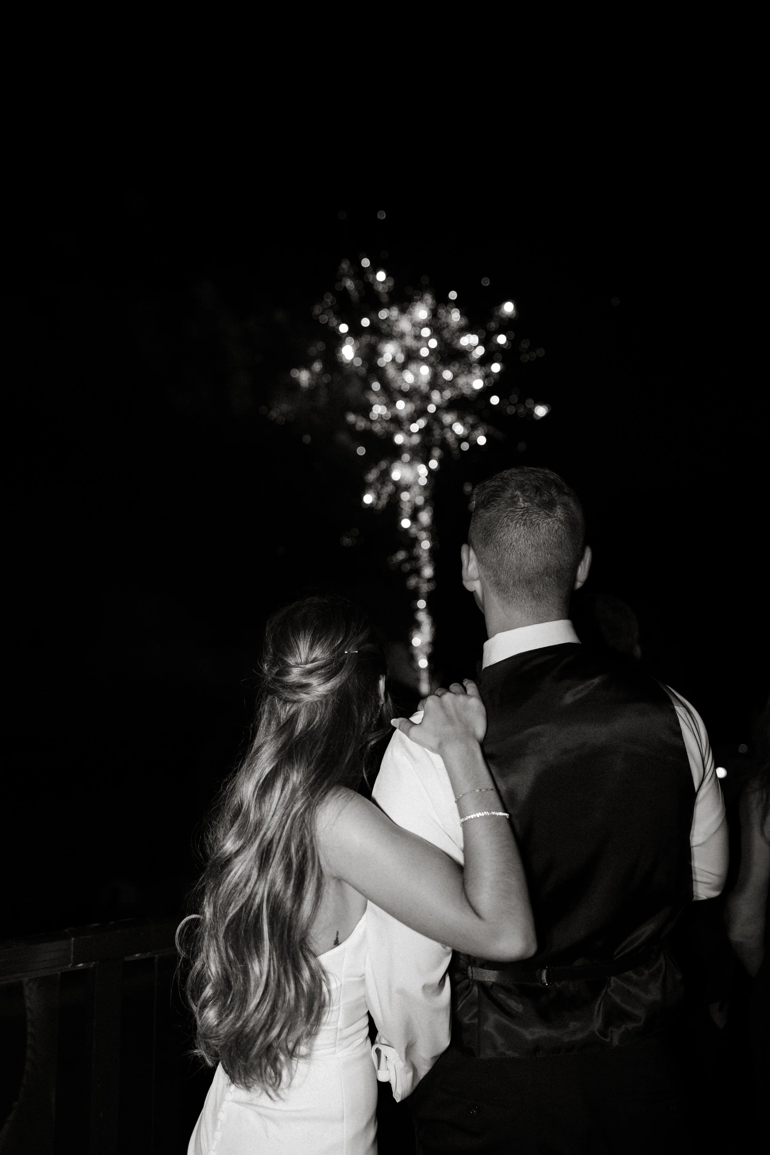 A couple watching fireworks at night, with the woman having long wavy hair and the man in a vest, illuminated by the fireworks.
