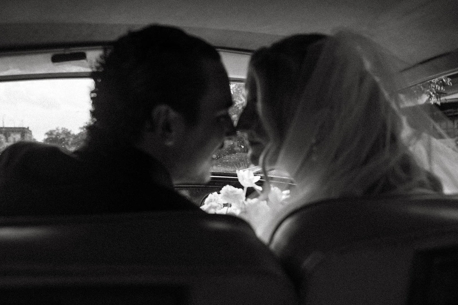 A black and white photo of a couple sitting close together in a car, leaning in towards each other with their foreheads touching, smiling, with flowers on the dashboard.