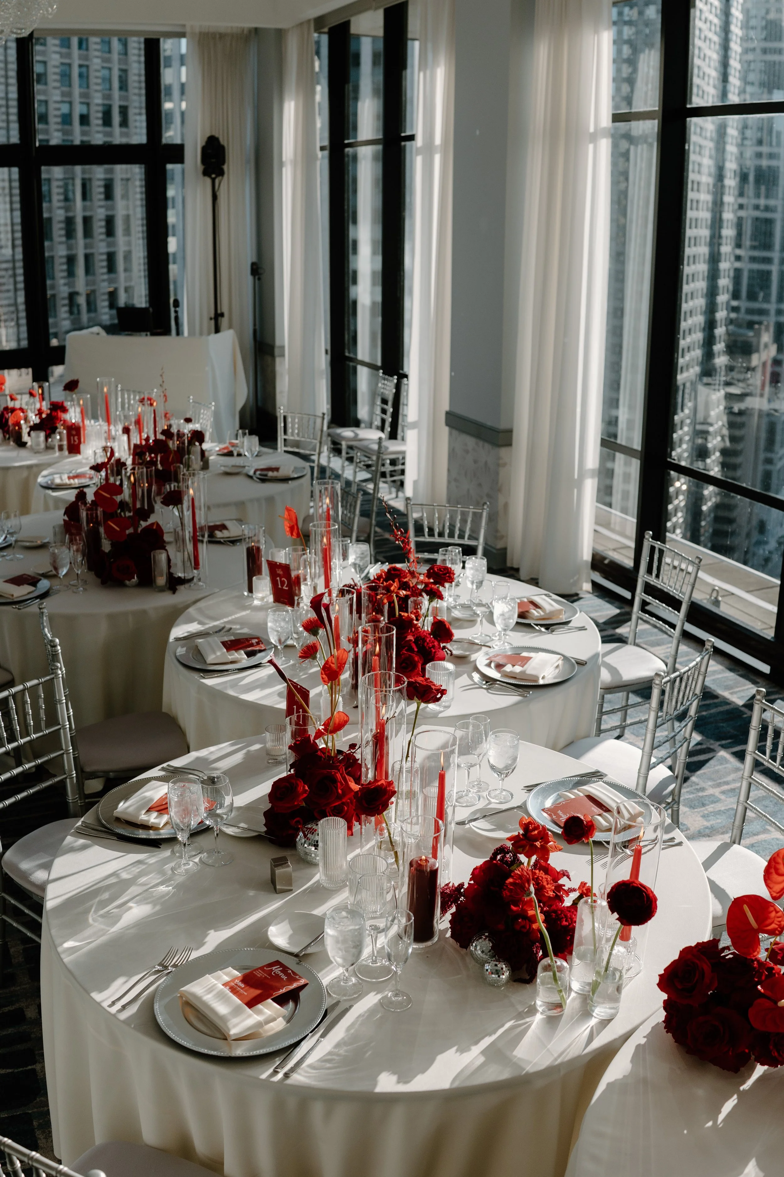 Elegant banquet table with white tablecloths, red floral centerpieces, assorted candles, and place settings with napkins, glasses, and cutlery, set in a high-rise room with large windows overlooking city buildings.