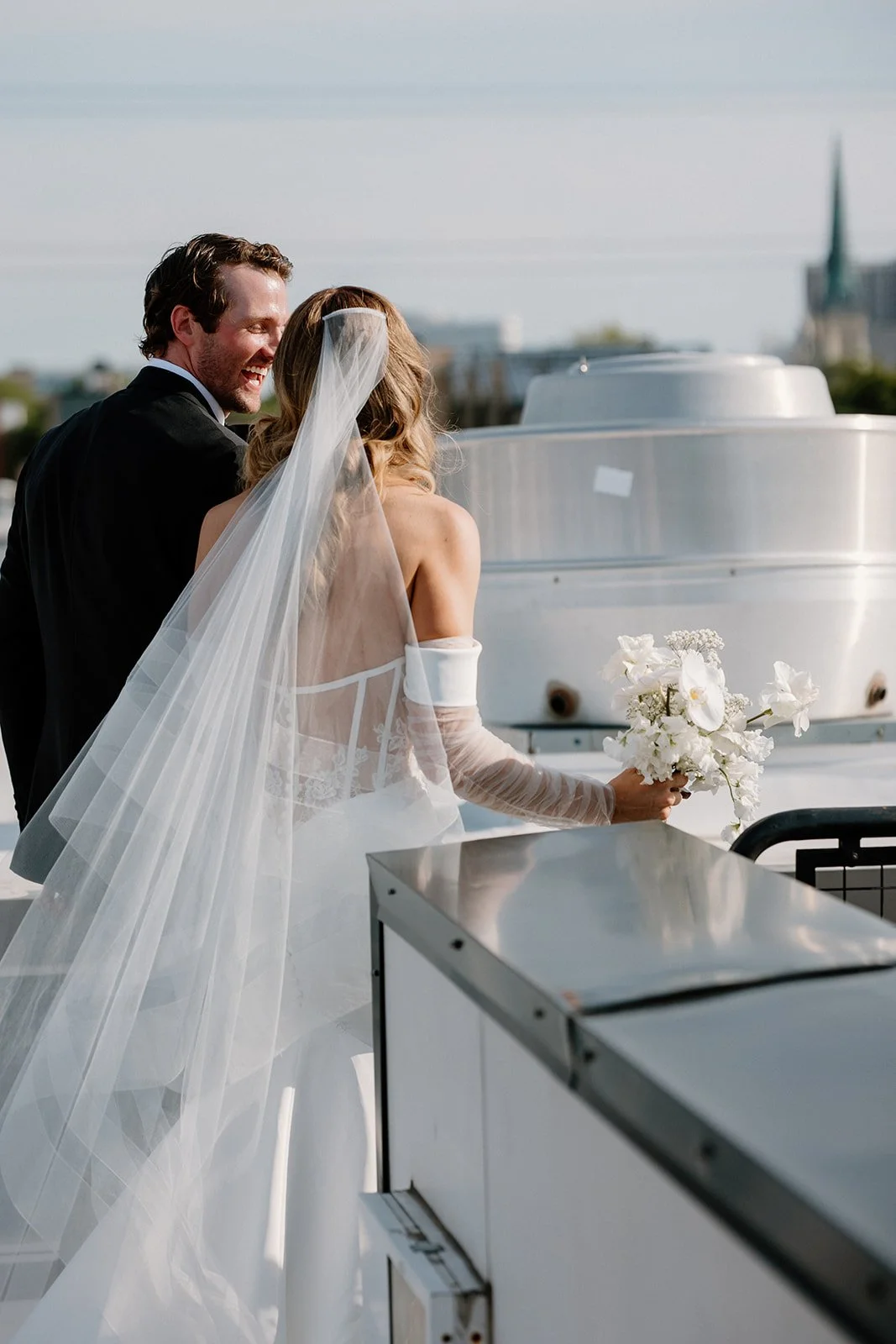 Bride and groom on a rooftop, the bride holding a bouquet of white flowers, both smiling and looking at each other, with a city skyline in the background.