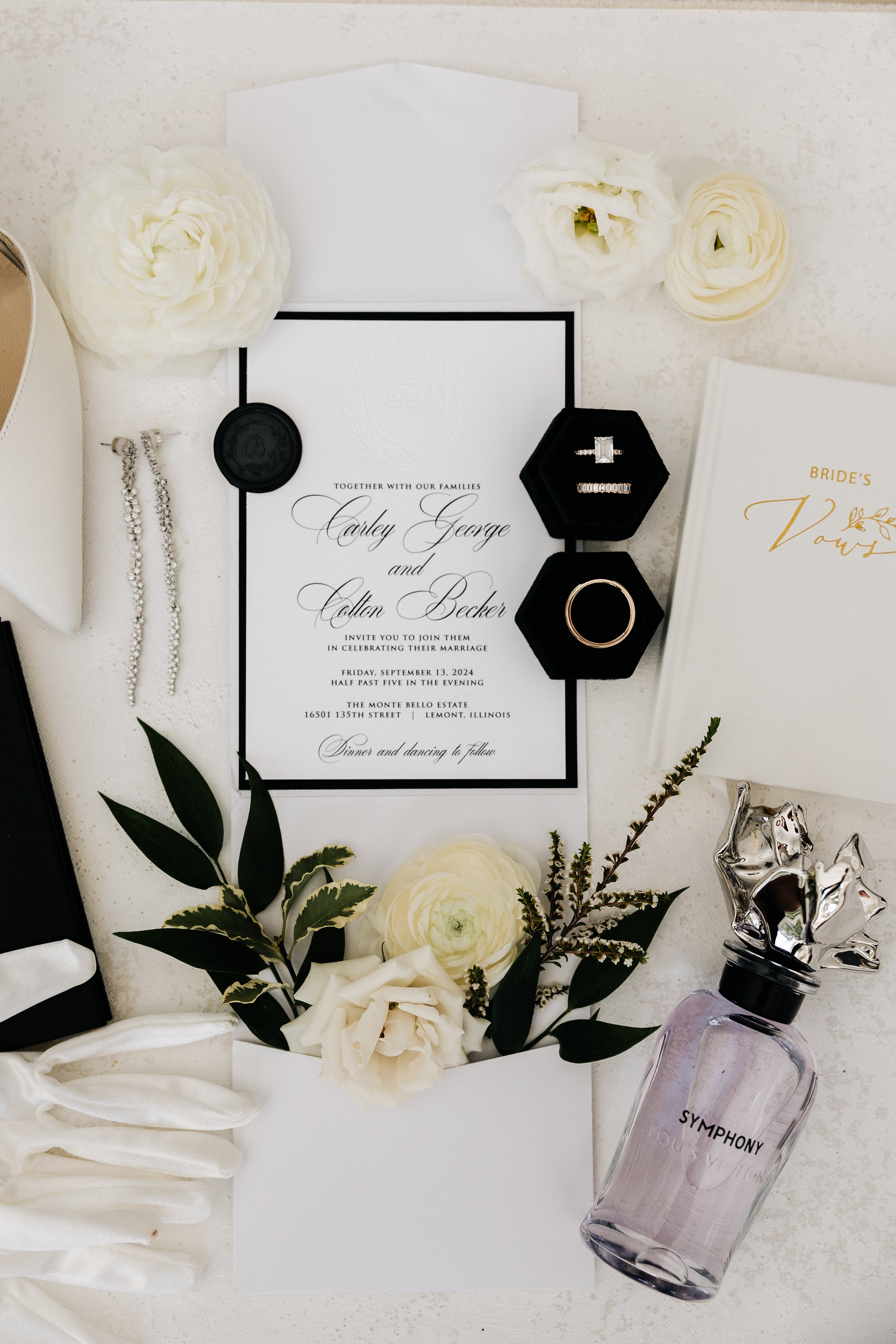 Wedding invitation surrounded by white flowers, jewelry, perfume, and a book titled 'Bride's Vows' on a white surface.